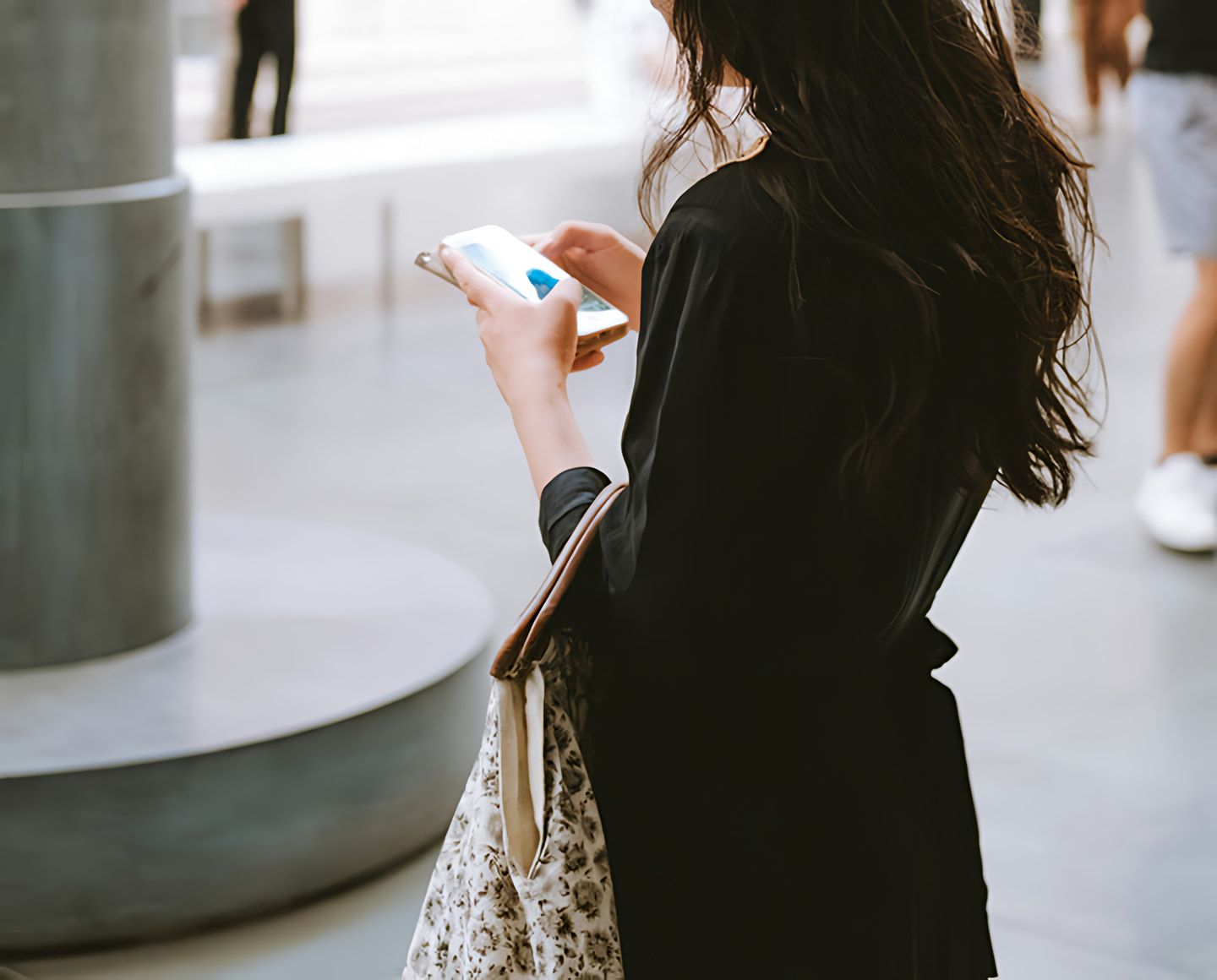 Woman in black clothing looking at a smartphone while carrying a patterned tote bag.