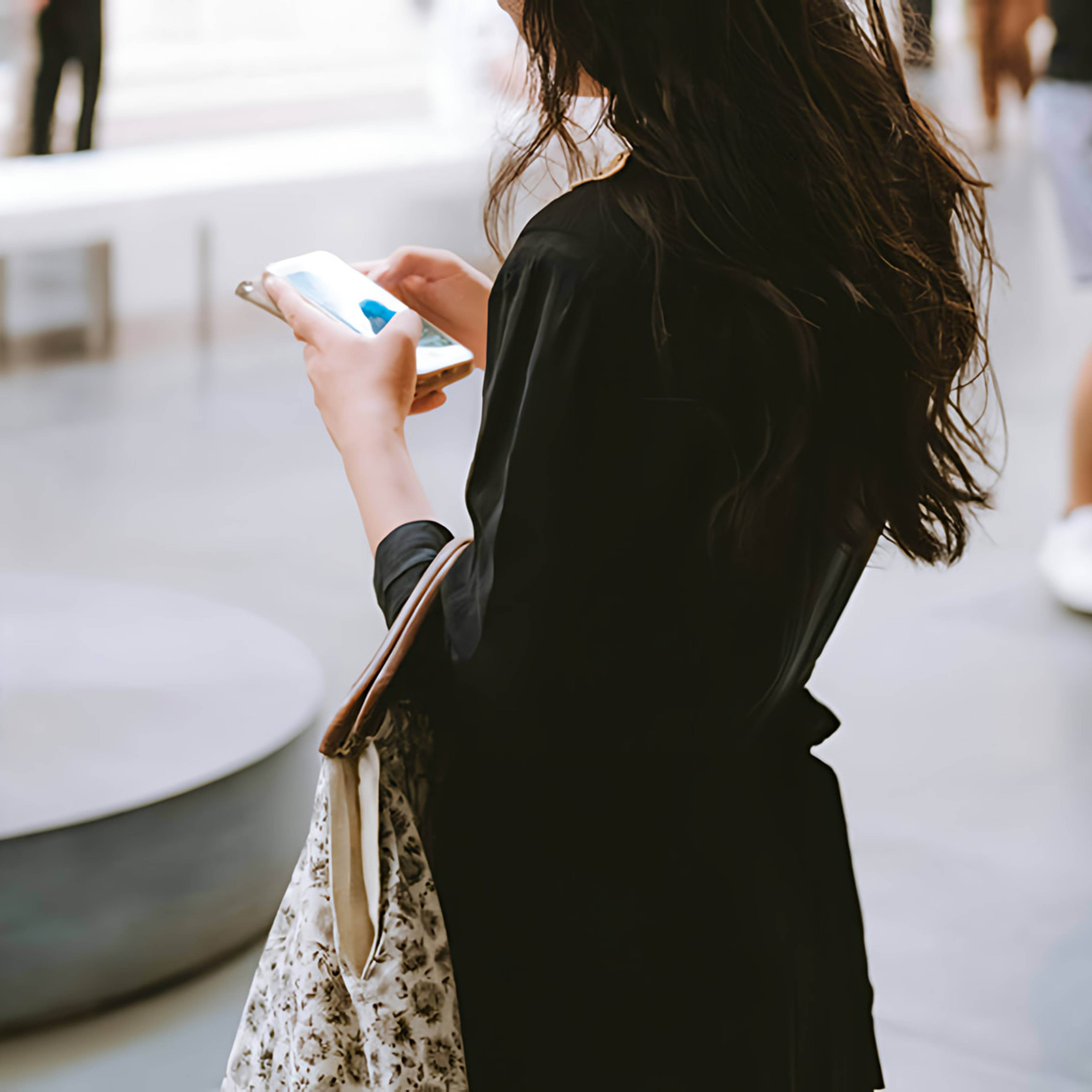 Woman in black clothing looking at a smartphone while carrying a patterned tote bag.