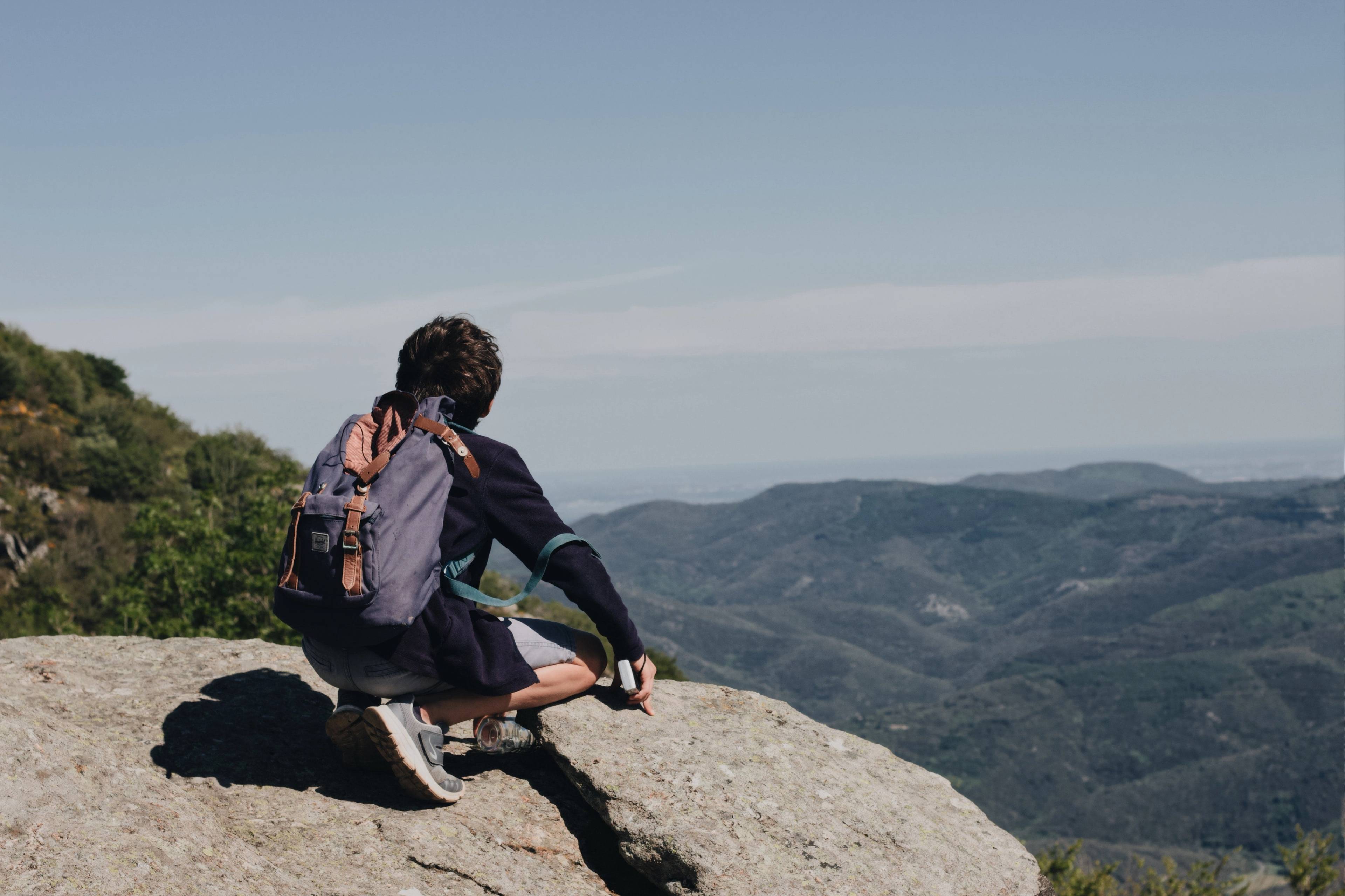 Hiker with a backpack sitting on a rocky overlook above forested mountains.