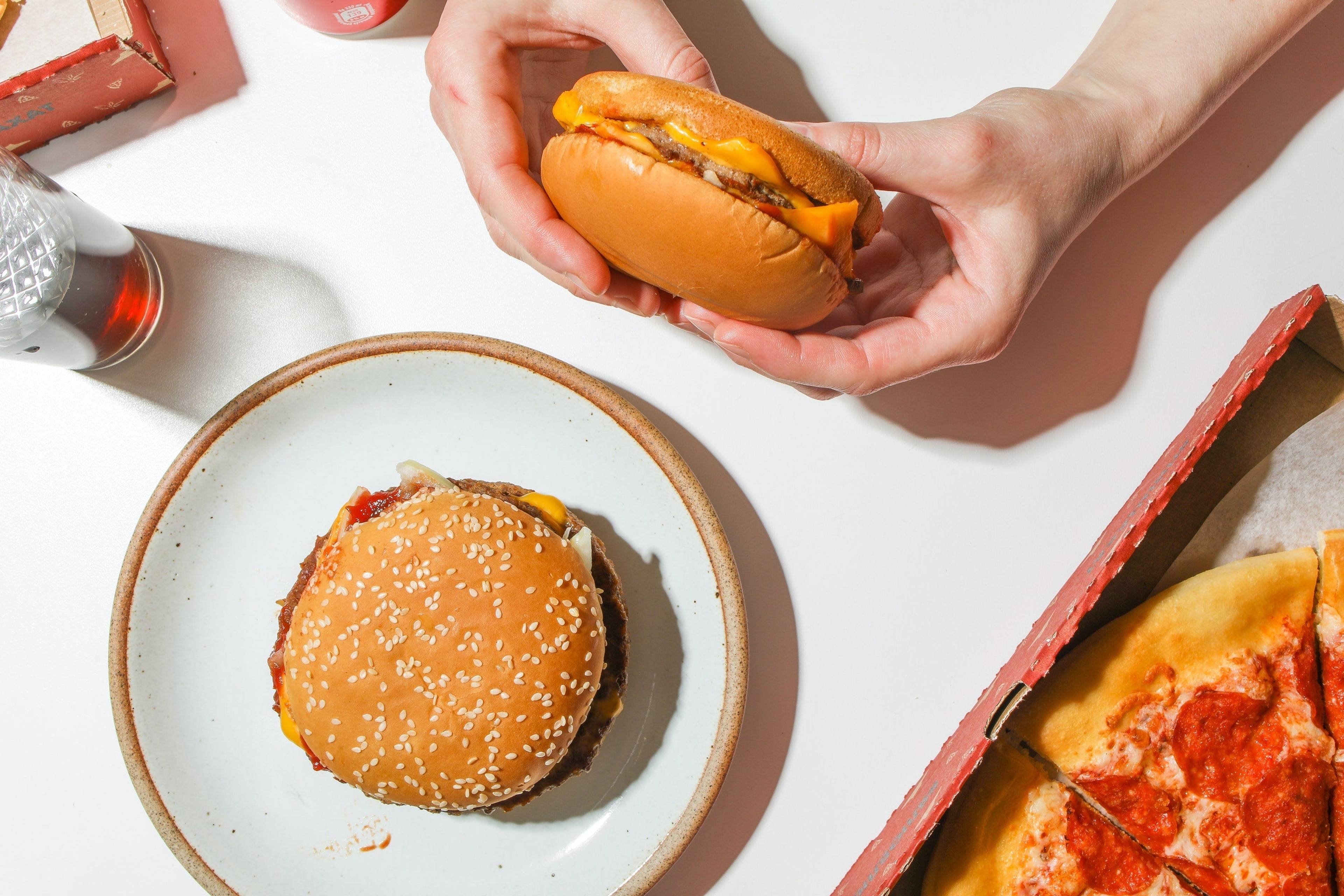 Hands holding a cheeseburger above a table with a sesame-seed burger, pizza slices, and a drink.