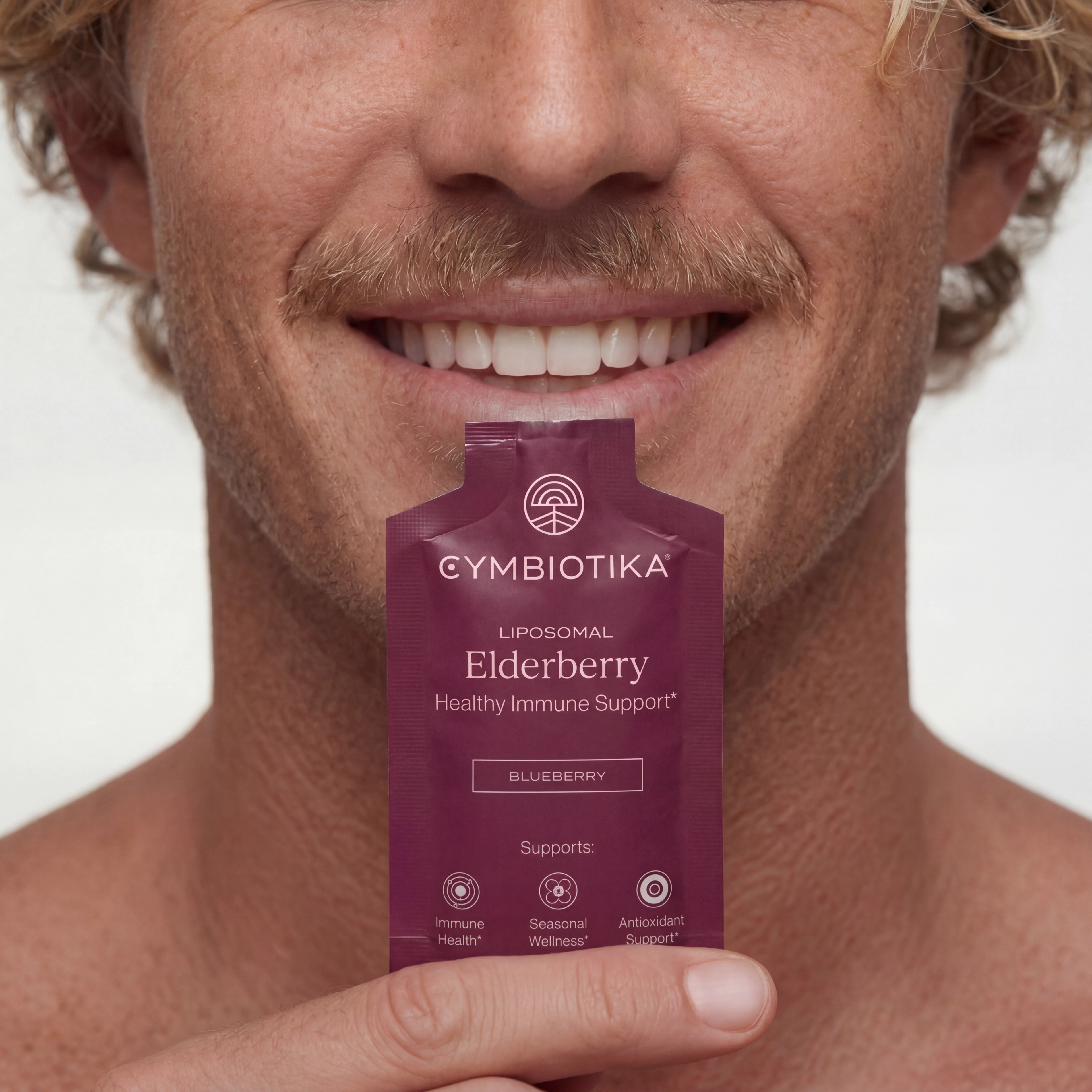 Smiling man holds a Cymbiotika Liposomal Elderberry packet just below his mouth.