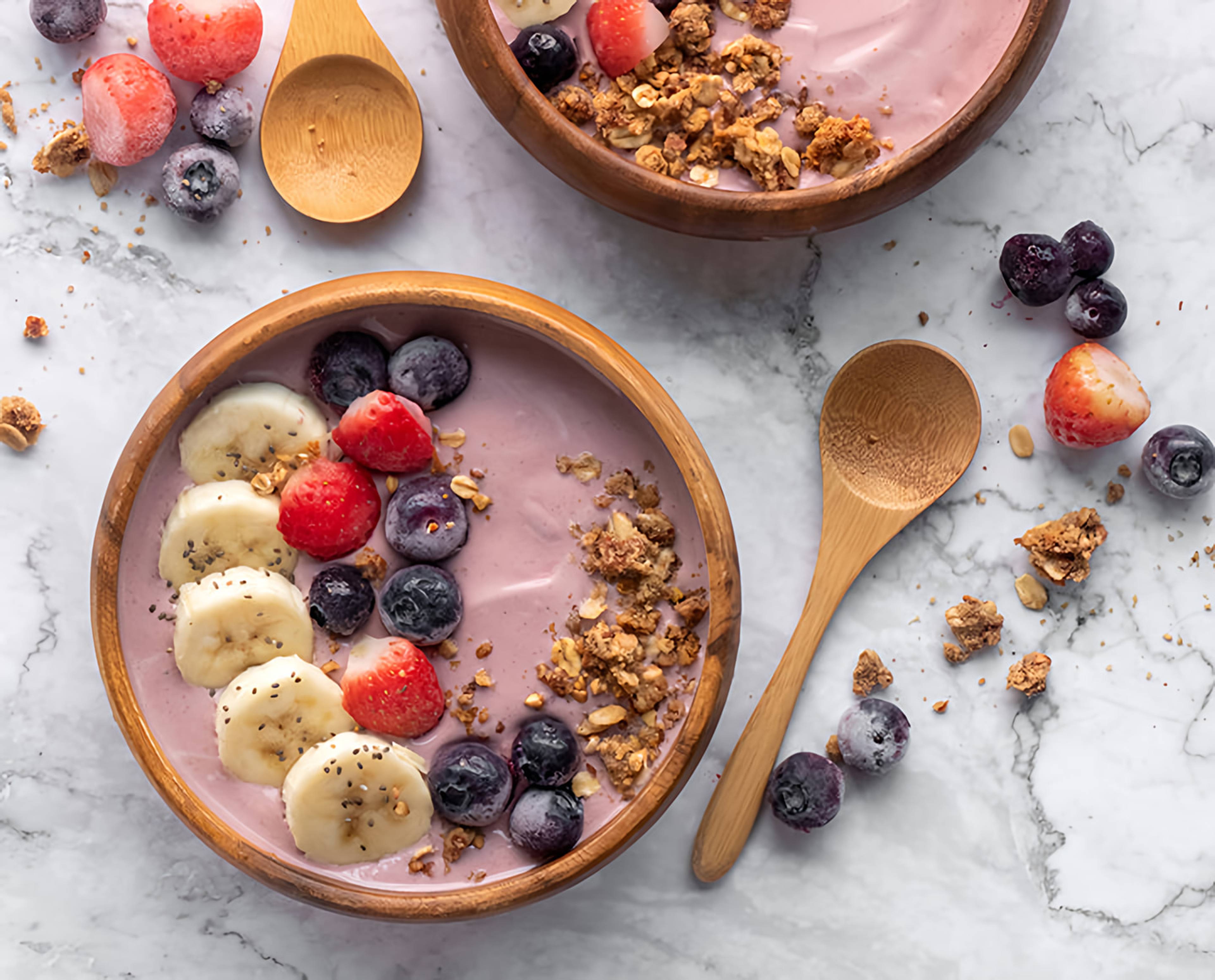 Two pink smoothie bowls topped with banana, strawberries, blueberries, granola, and chia seeds beside wooden spoons.