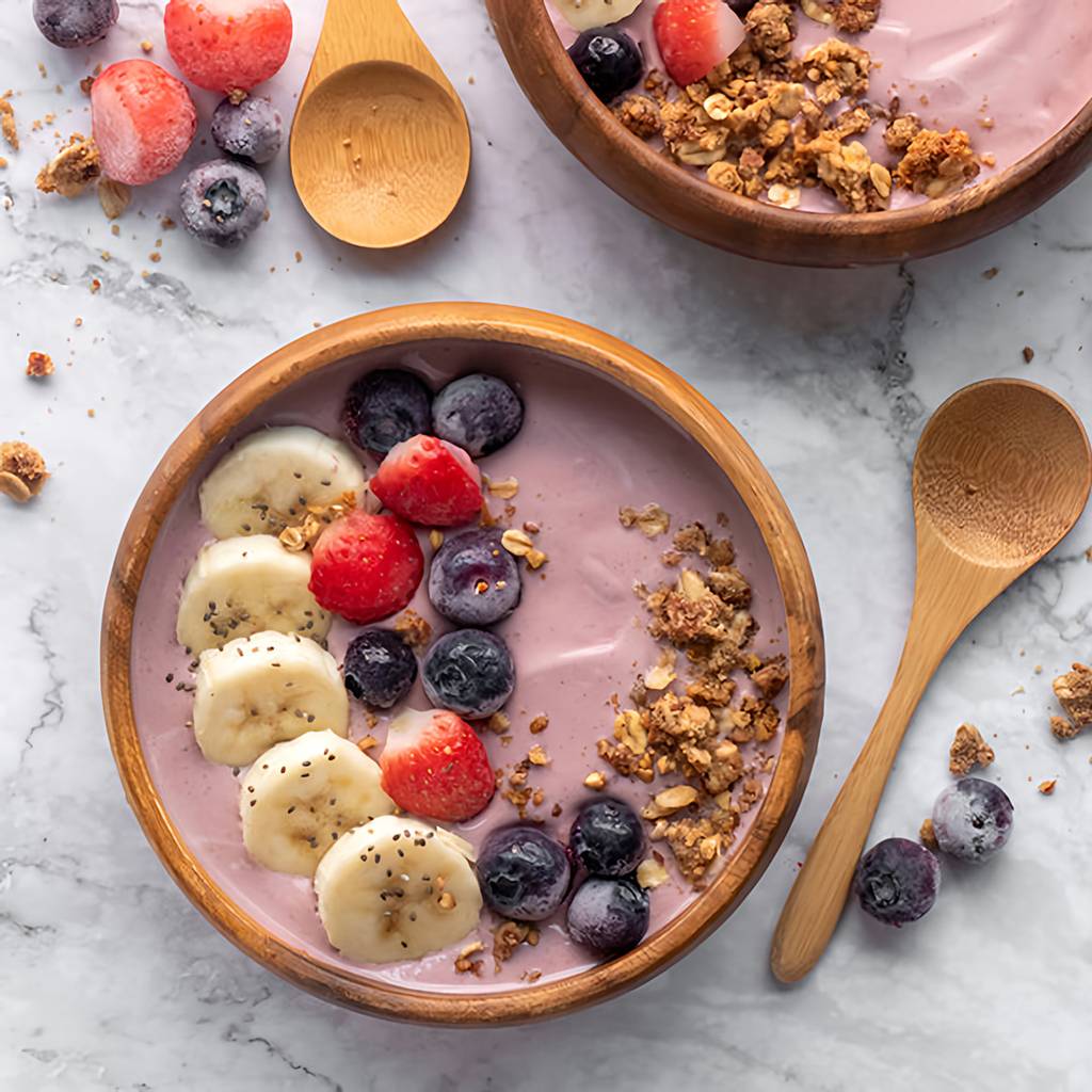 Two pink smoothie bowls topped with banana, strawberries, blueberries, granola, and chia seeds beside wooden spoons.