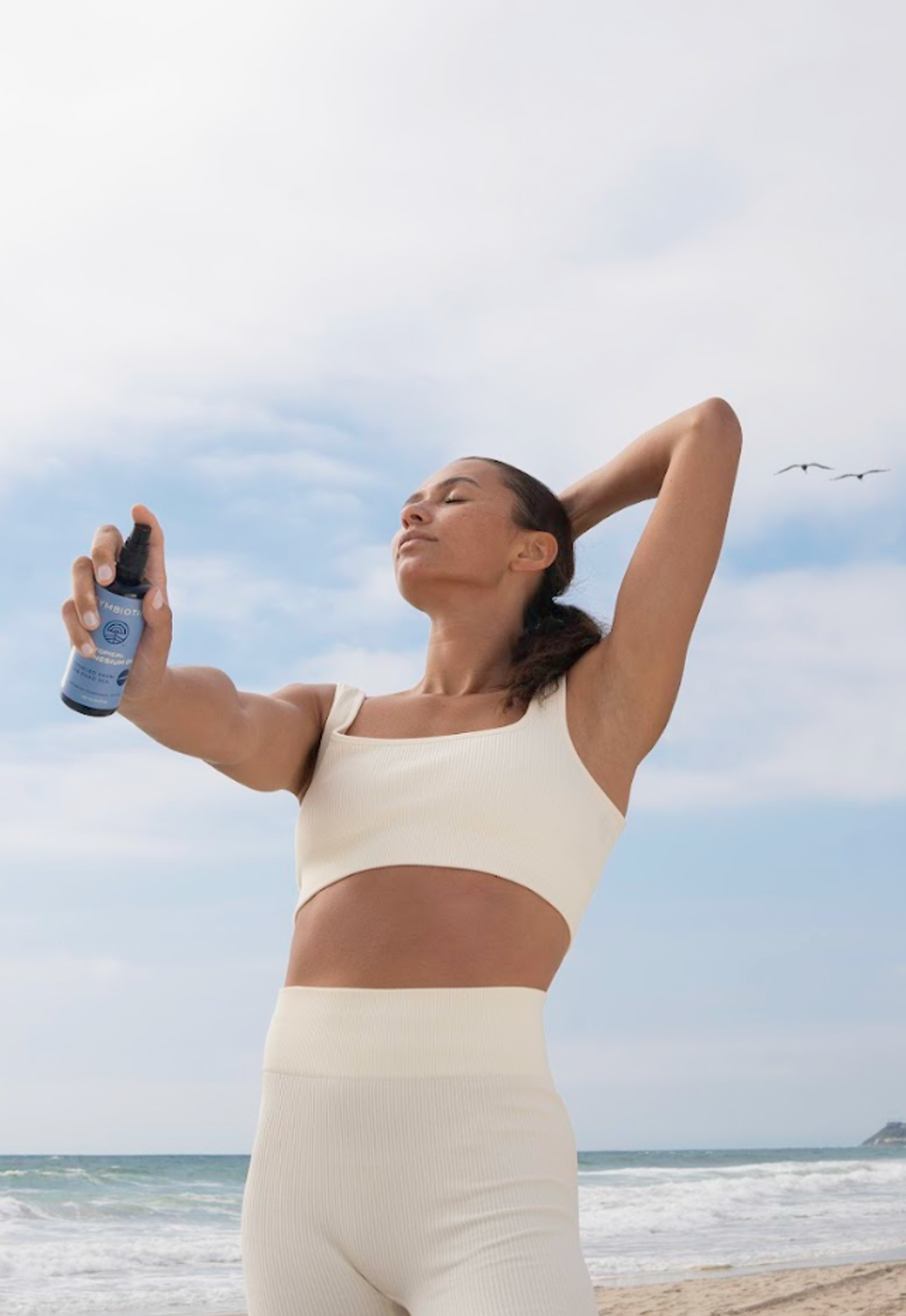Woman in cream activewear holds a blue spray bottle on a beach with her eyes closed.