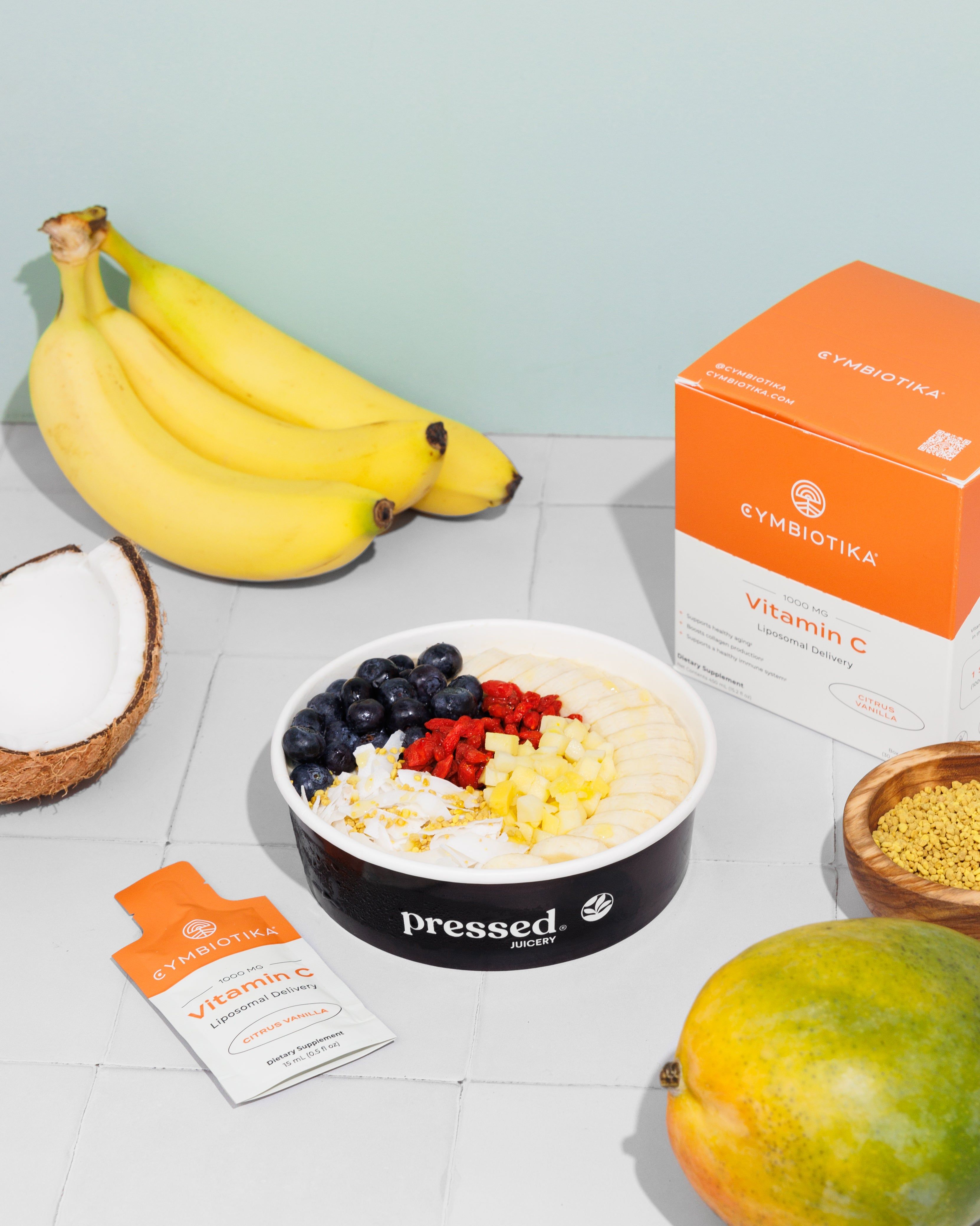 Pressed Juicery fruit bowl with bananas, coconut, blueberries, raspberries, mango, and Cymbiotika Liposomal Vitamin C box and packet.