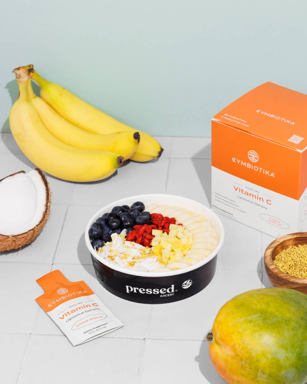 Pressed Juicery fruit bowl with bananas, coconut, blueberries, raspberries, mango, and Cymbiotika Liposomal Vitamin C box and packet.