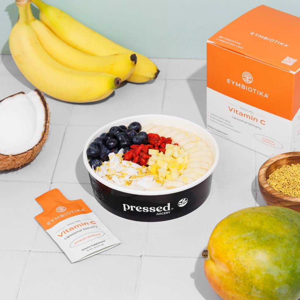 Pressed Juicery fruit bowl with bananas, coconut, blueberries, raspberries, mango, and Cymbiotika Liposomal Vitamin C box and packet.