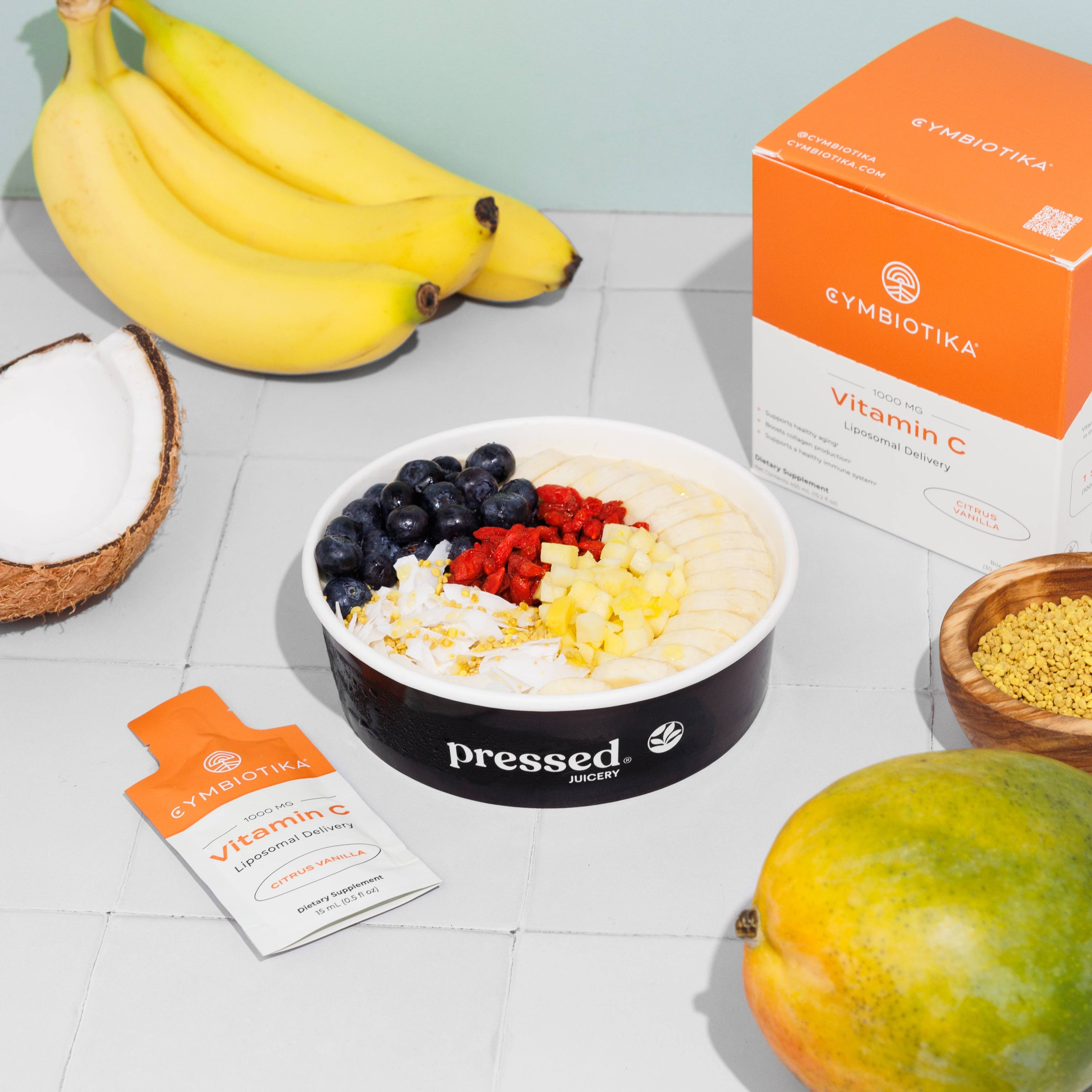 Pressed Juicery fruit bowl with bananas, coconut, blueberries, raspberries, mango, and Cymbiotika Liposomal Vitamin C box and packet.