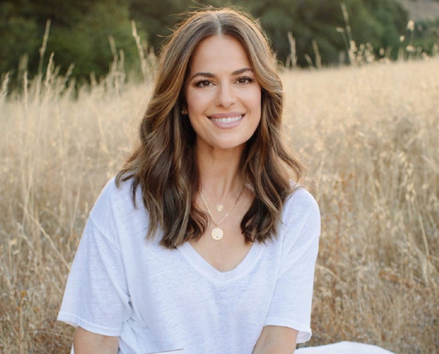 Smiling woman in a white shirt seated in a sunlit field of dry grass.