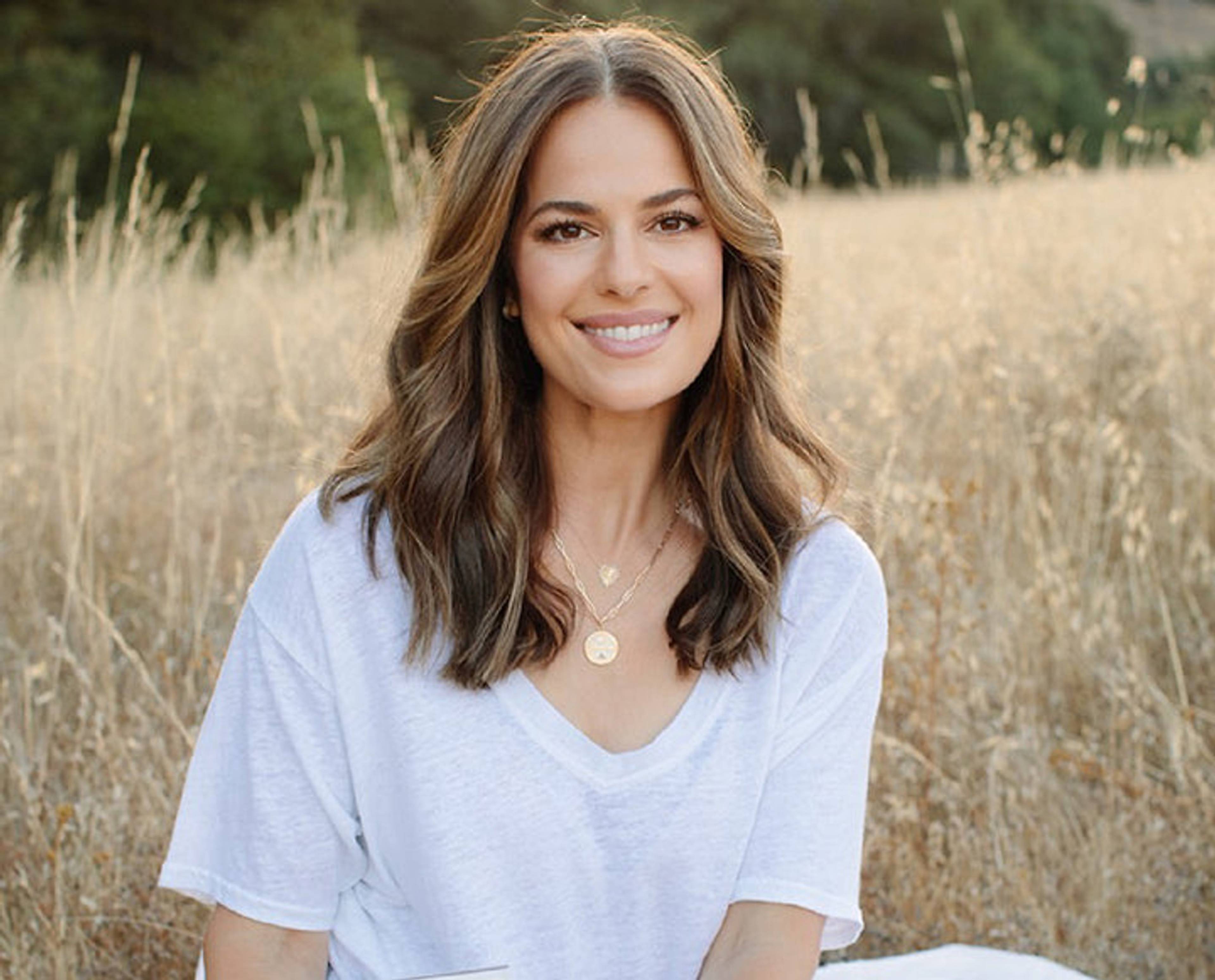 Smiling woman in a white shirt seated in a sunlit field of dry grass.