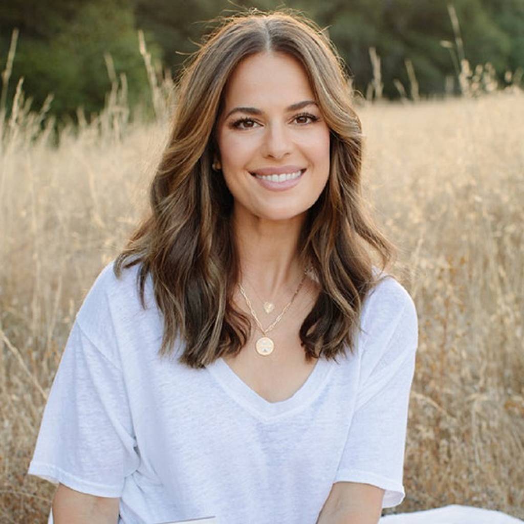 Smiling woman in a white shirt seated in a sunlit field of dry grass.