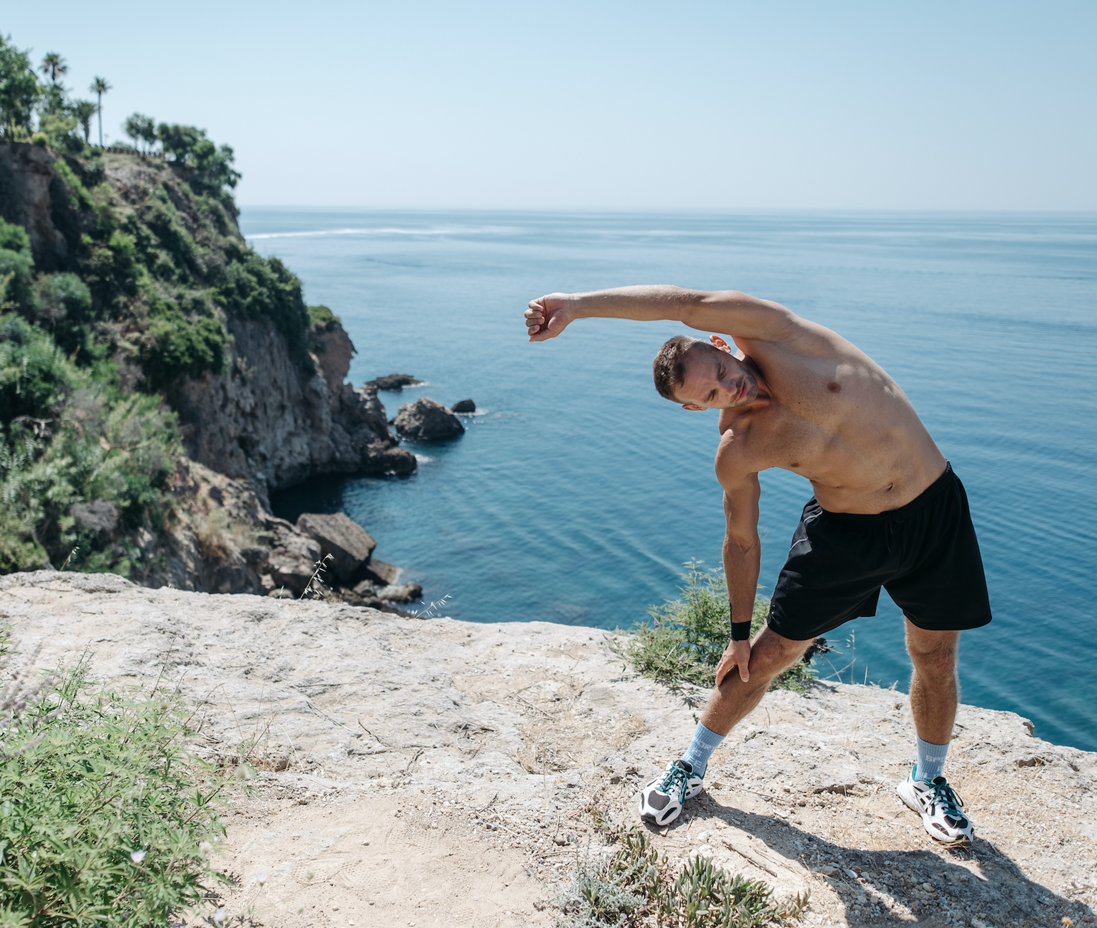 Shirtless man stretching on a rocky coastal cliff above the ocean.
