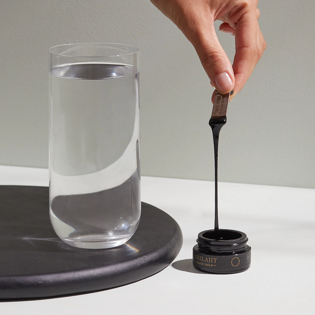 A hand lifts a strand of dark resin from an open jar labeled “Shilajit,” beside a glass of water.