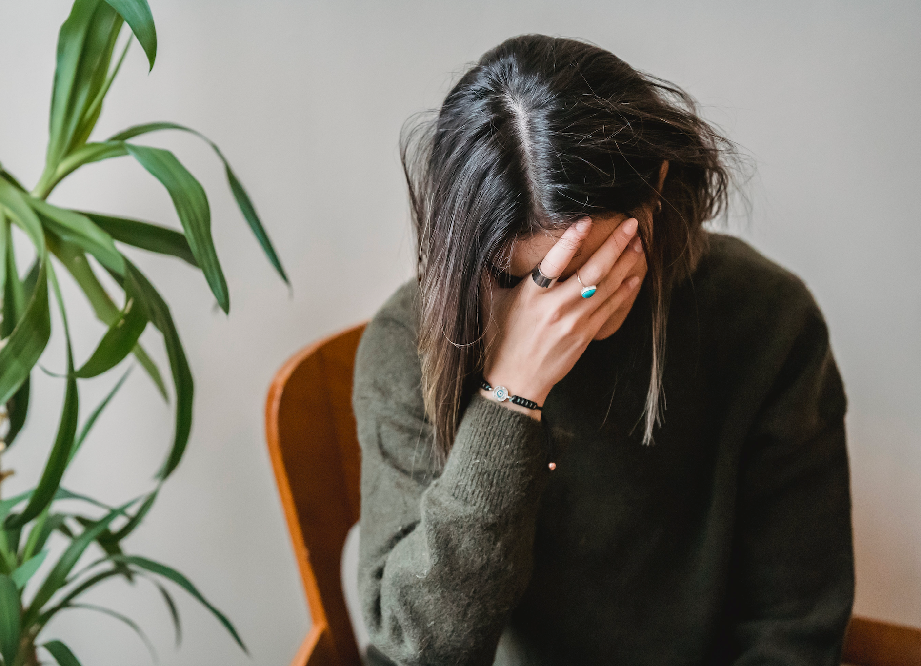Person in a dark sweater sits with their face covered by one hand beside a leafy houseplant.