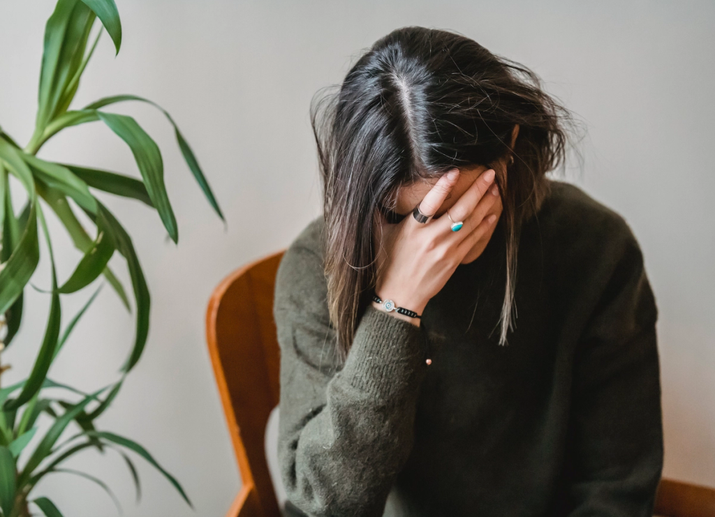 Person in a dark sweater sits with their face covered by one hand beside a leafy houseplant.