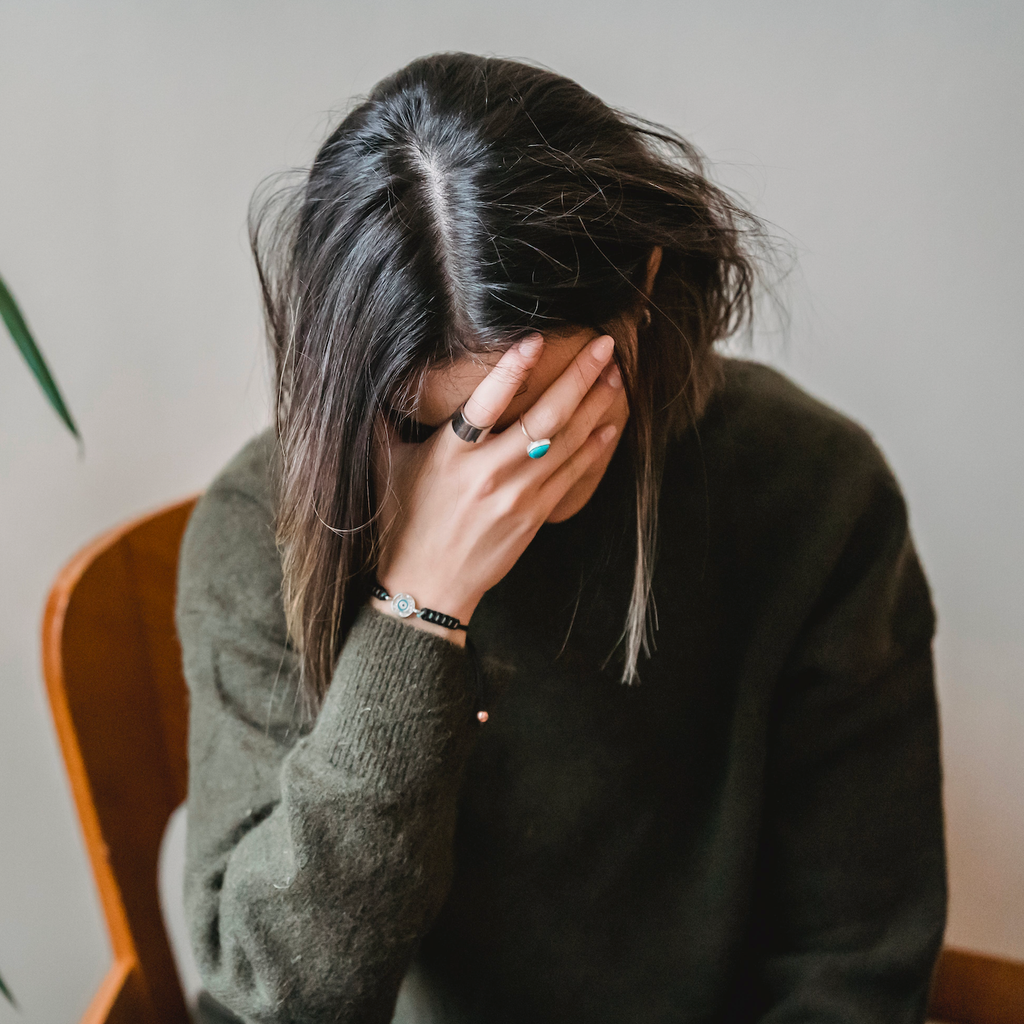 Person in a dark sweater sits with their face covered by one hand beside a leafy houseplant.