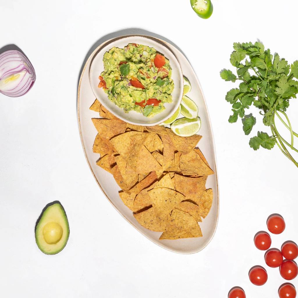 Bowl of guacamole with tortilla chips, lime wedges, jalapeño, cilantro, avocado, red onion, and cherry tomatoes.