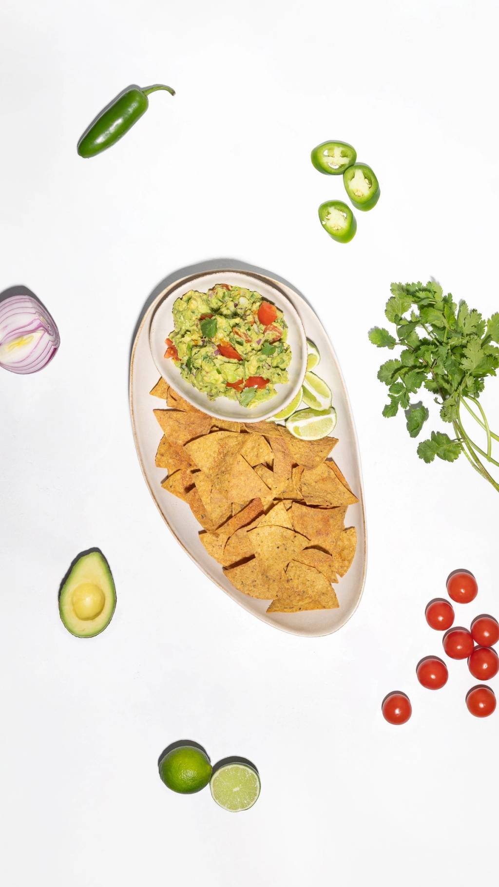 Bowl of guacamole with tortilla chips, lime wedges, jalapeño, cilantro, avocado, red onion, and cherry tomatoes.