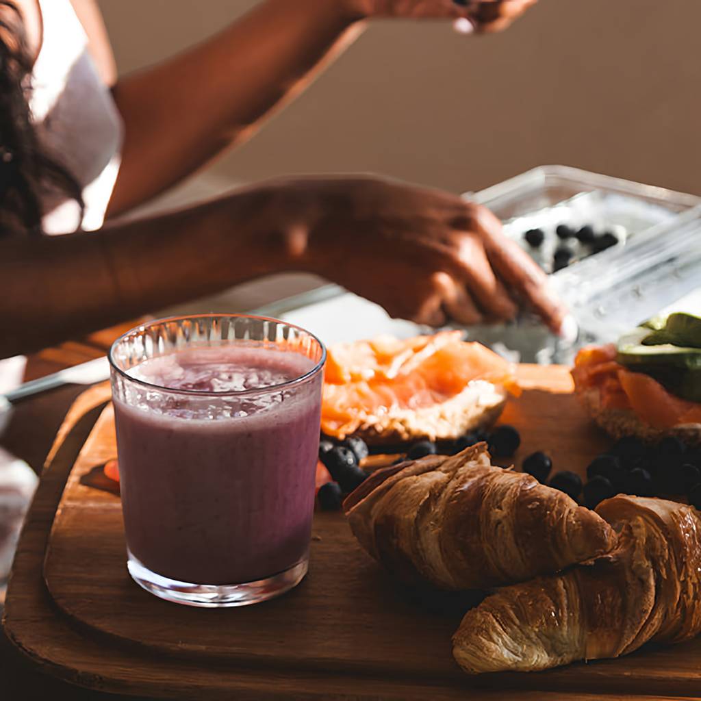 Person reaching for berries beside a pink smoothie, croissants, smoked salmon, and greens on a wooden board.