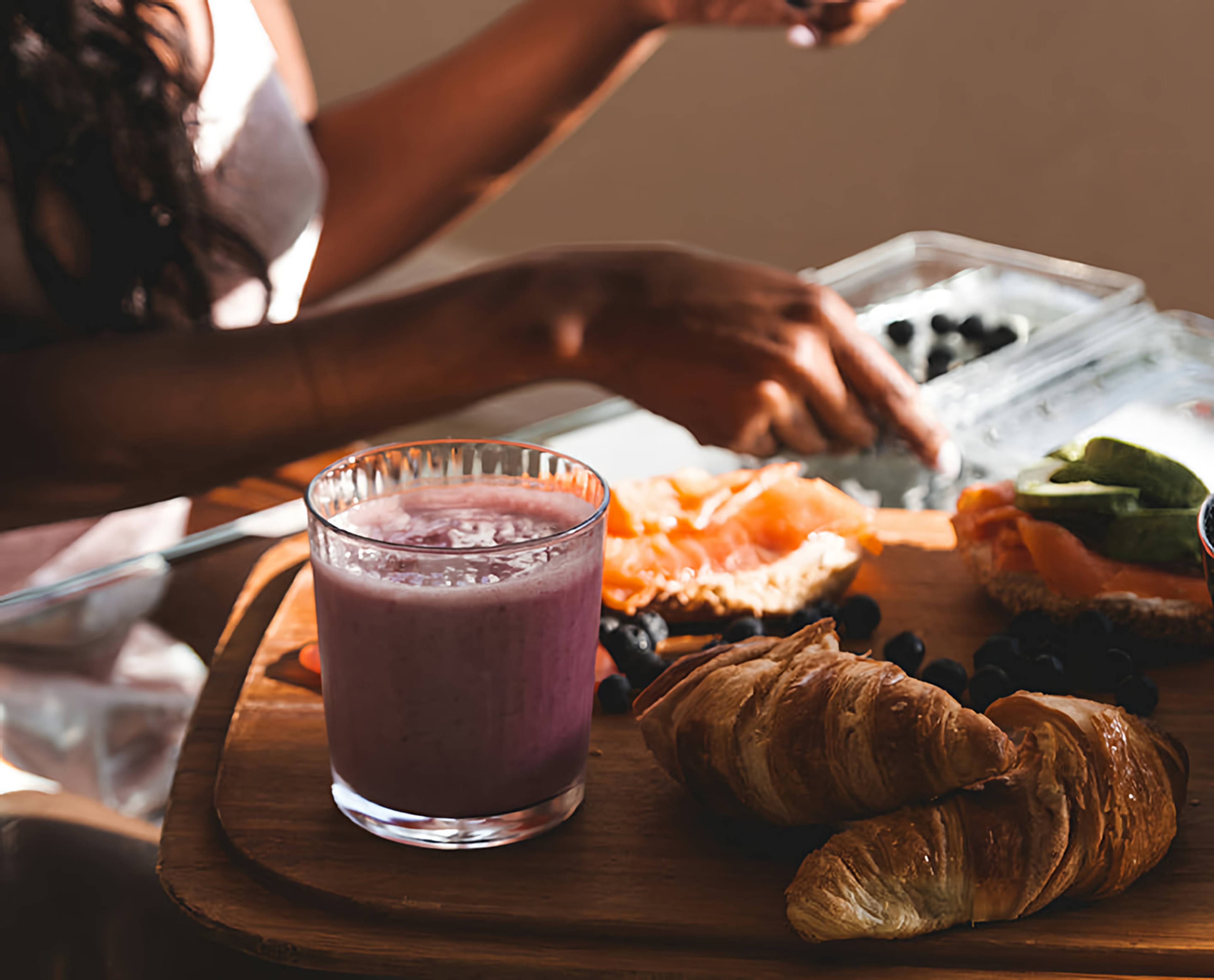 Person reaching for berries beside a pink smoothie, croissants, smoked salmon, and greens on a wooden board.