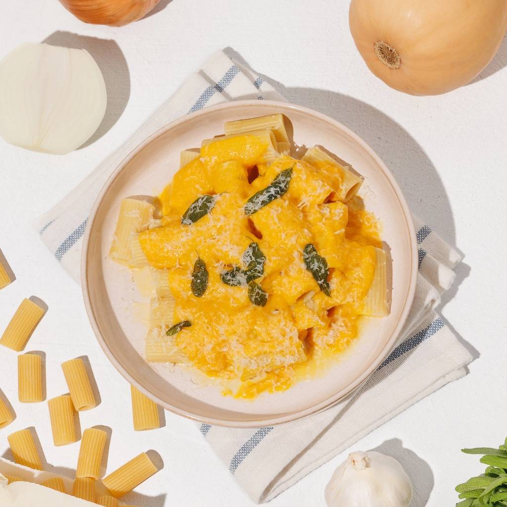 Rigatoni pasta with creamy orange squash sauce, sage, and grated cheese surrounded by squash, onion, garlic, and dry pasta.