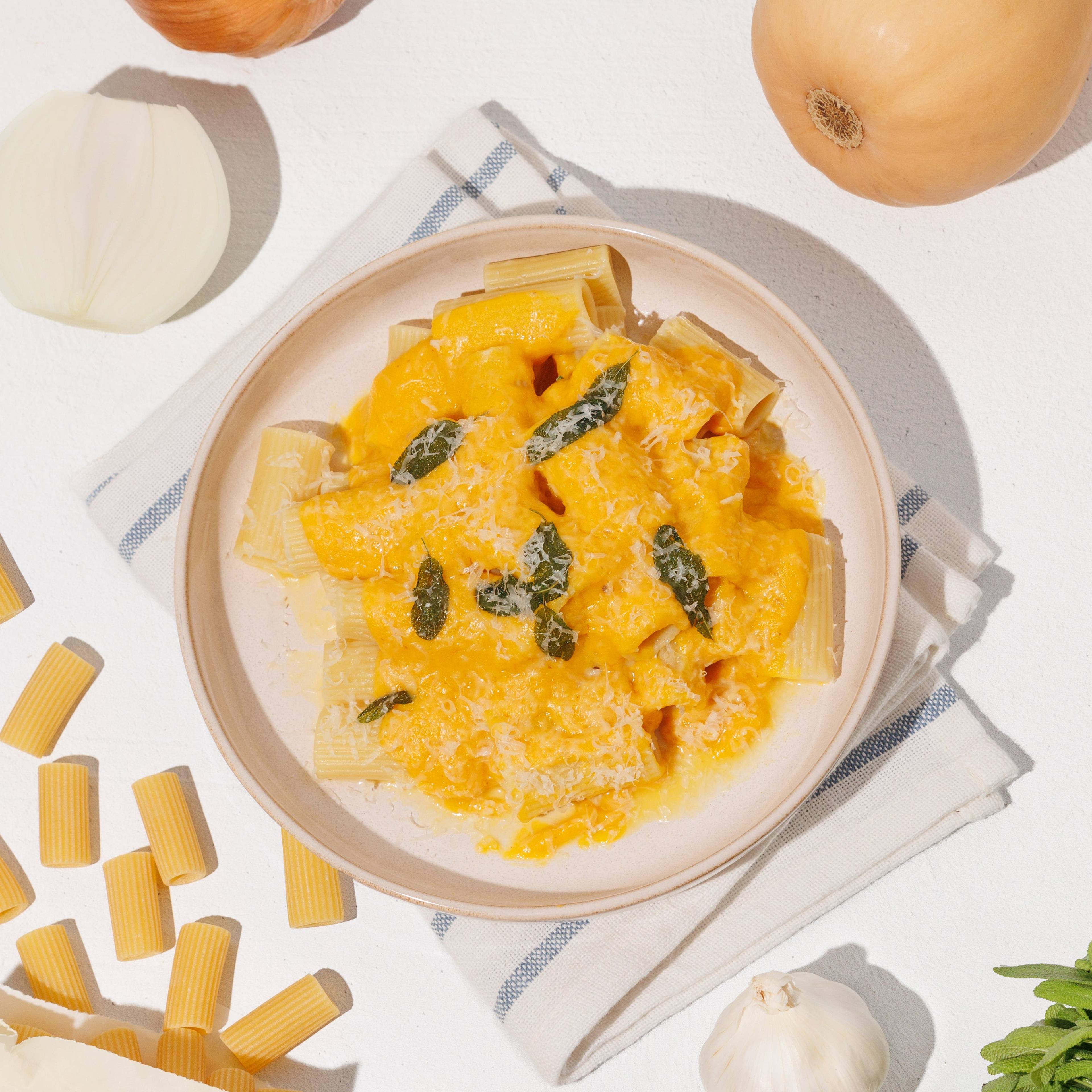 Rigatoni pasta with creamy orange squash sauce, sage, and grated cheese surrounded by squash, onion, garlic, and dry pasta.