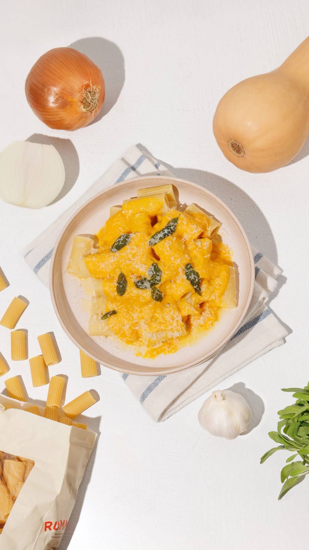 Rigatoni pasta with creamy orange squash sauce, sage, and grated cheese surrounded by squash, onion, garlic, and dry pasta.