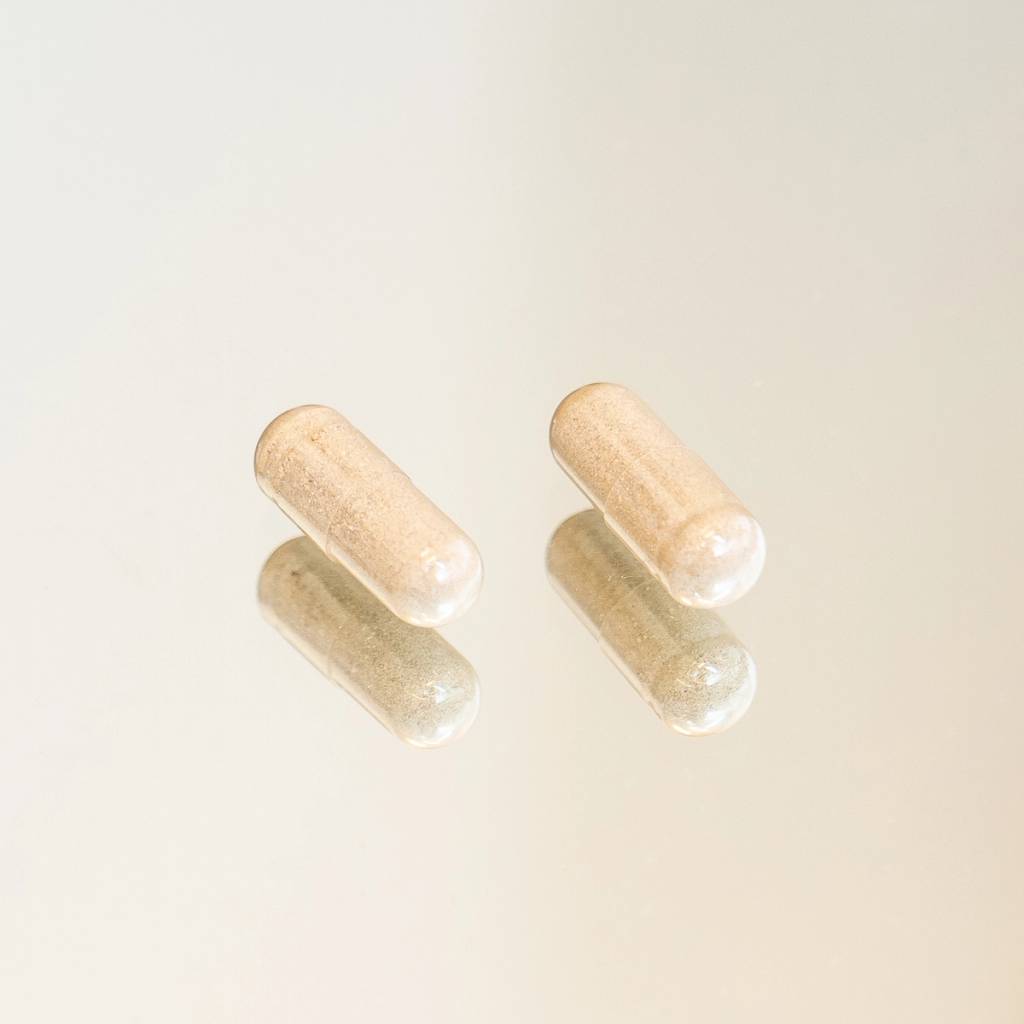 Two beige-filled supplement capsules with soft reflections on a pale background.