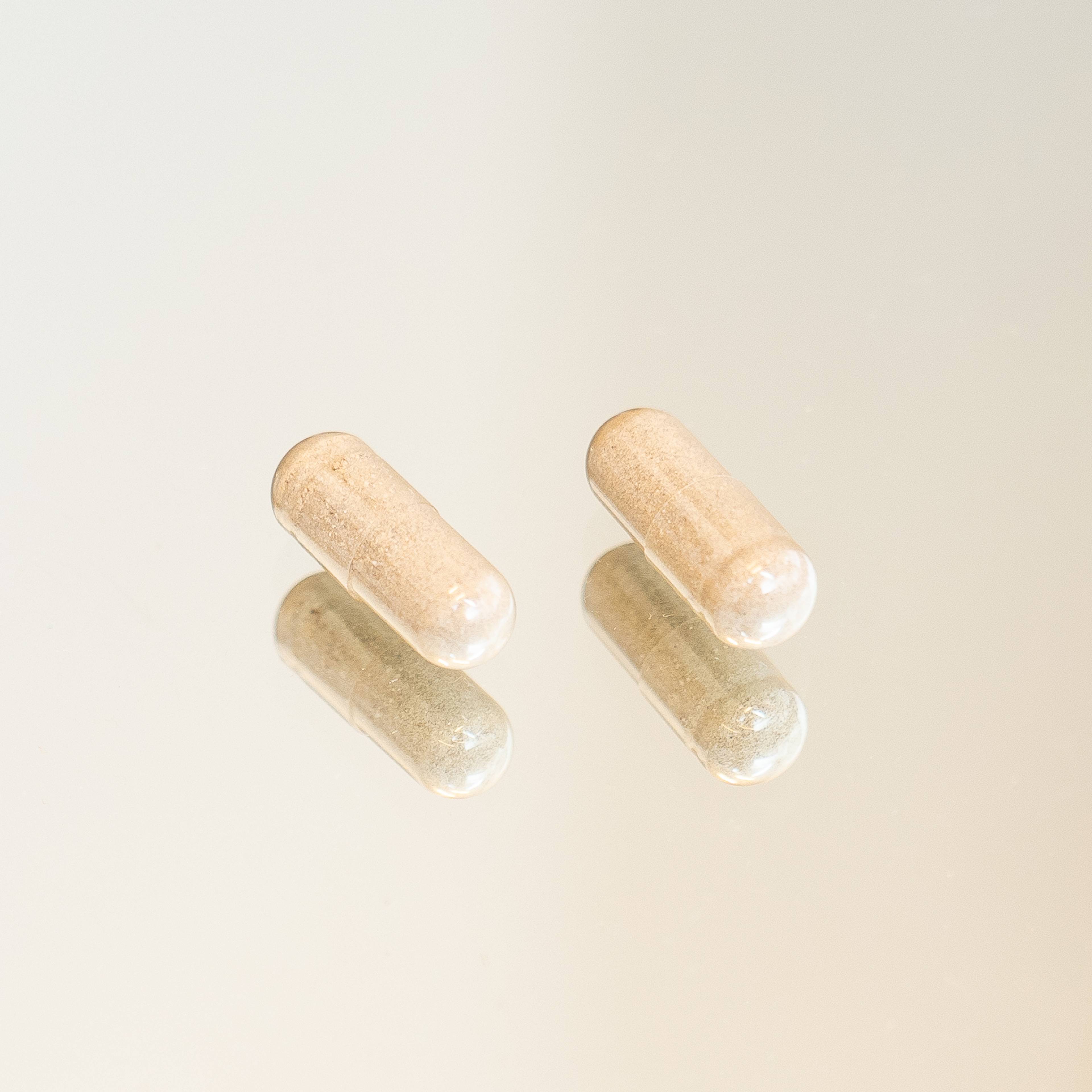 Two beige-filled supplement capsules with soft reflections on a pale background.