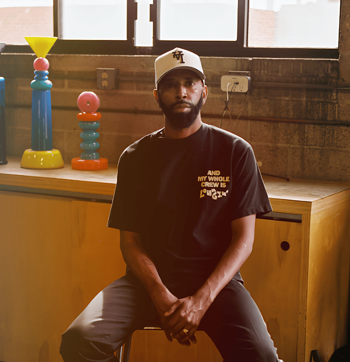 Bearded man in a baseball cap and black T-shirt seated indoors beside colorful stacked sculptures.
