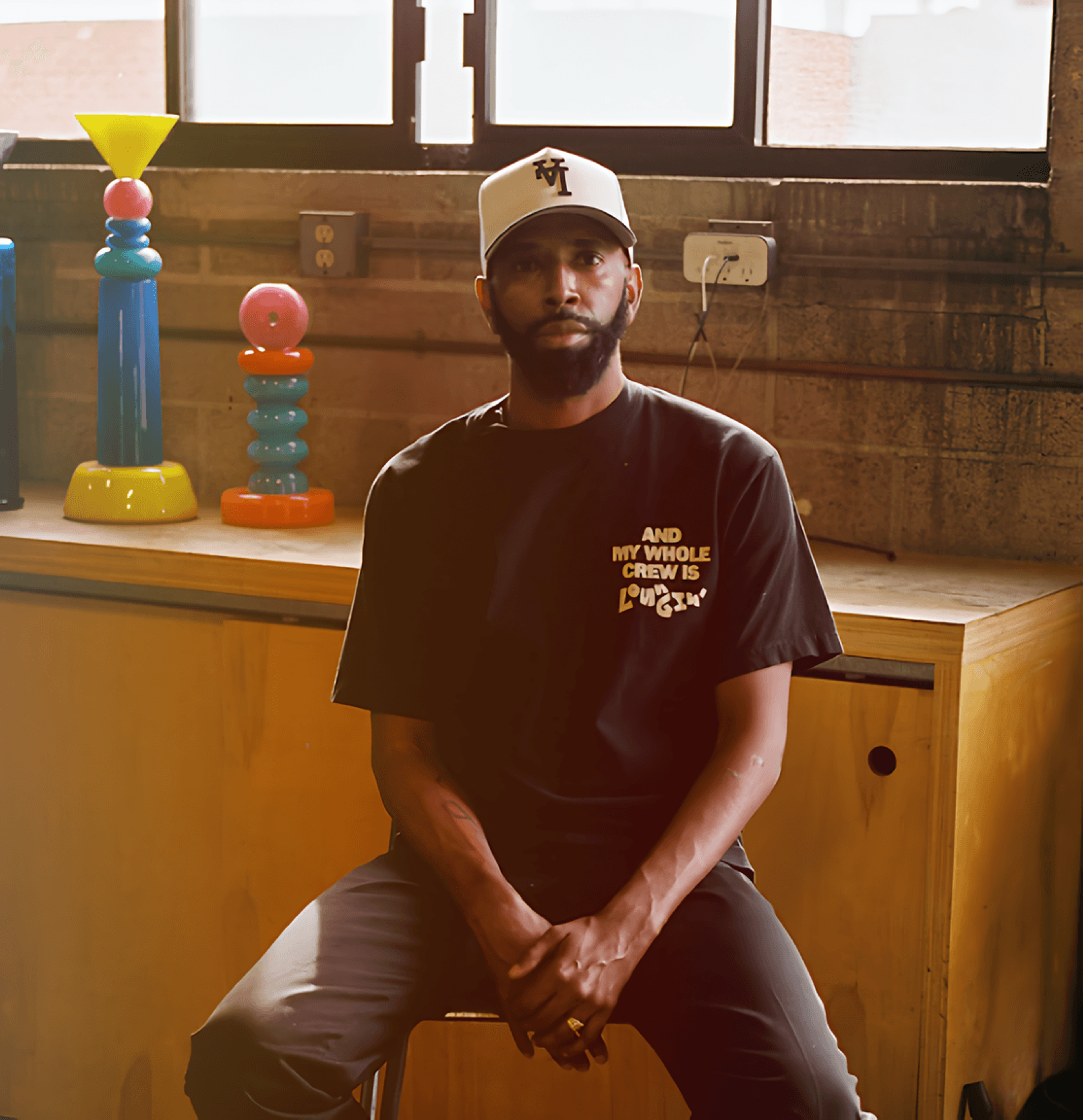 Bearded man in a baseball cap and black T-shirt seated indoors beside colorful stacked sculptures.