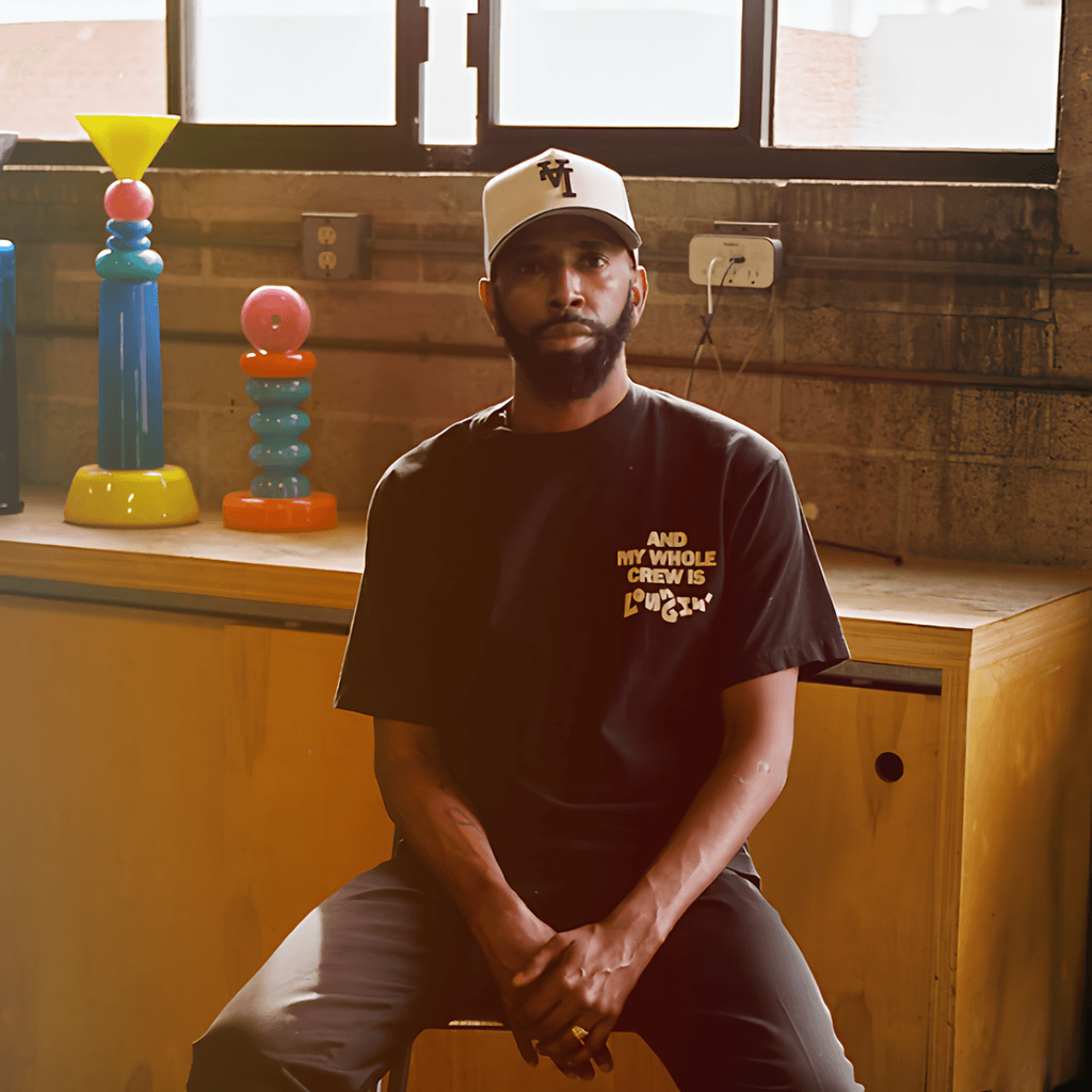 Bearded man in a baseball cap and black T-shirt seated indoors beside colorful stacked sculptures.