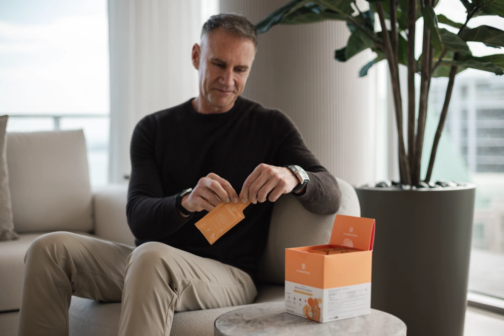 Man seated indoors opening an orange supplement packet beside an open orange box on a table.