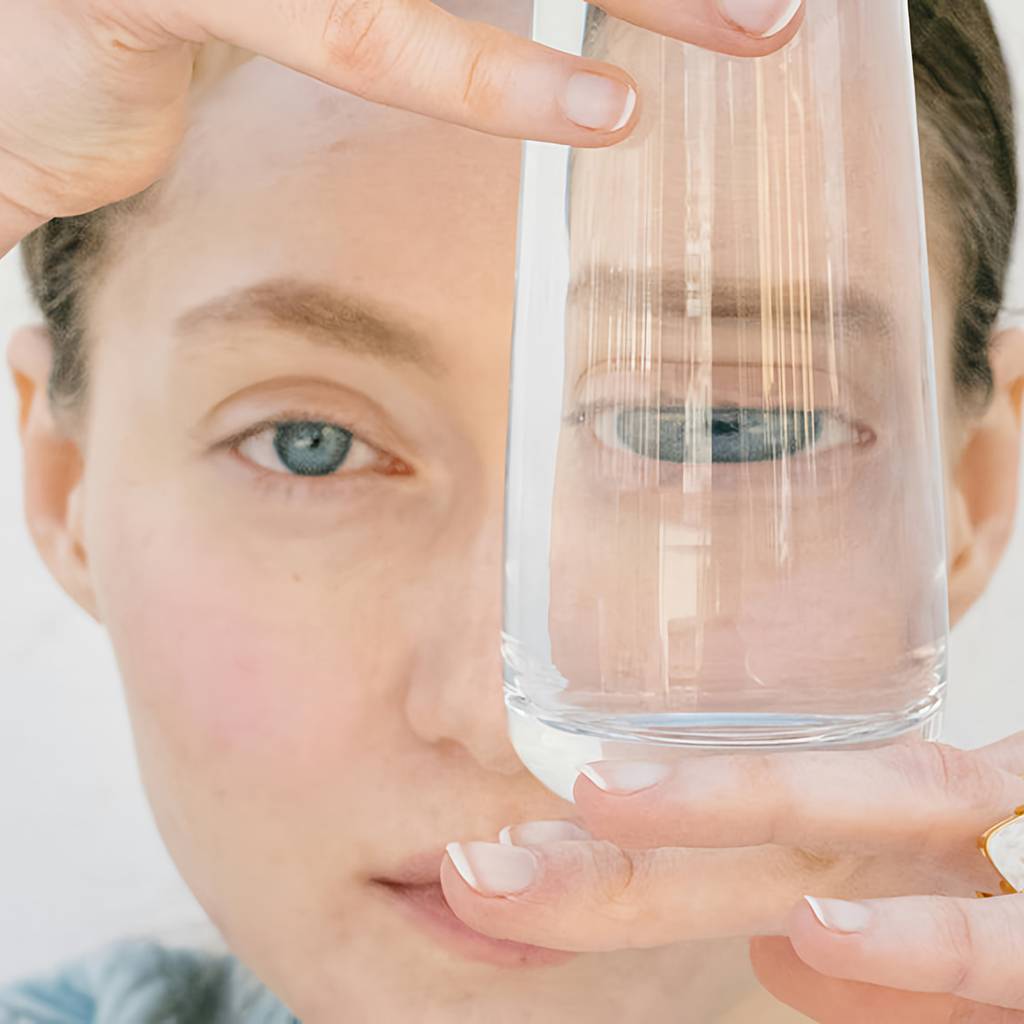 Close-up of a woman holding a clear glass of water in front of one eye.