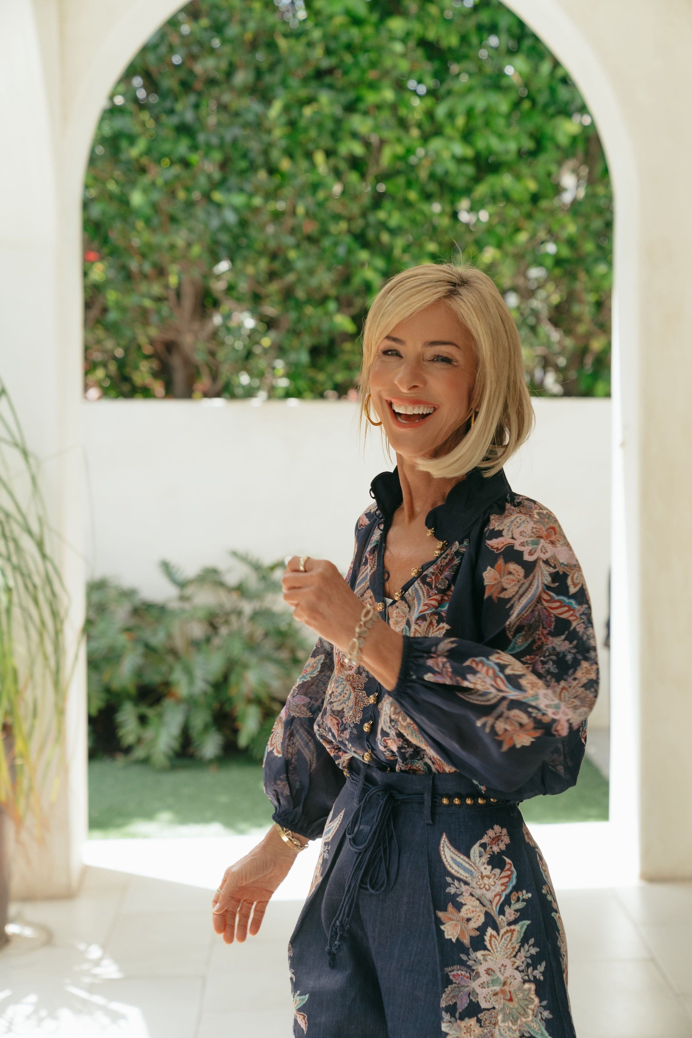 A smiling blonde woman in a dark floral-patterned outfit stands beside an arched opening with greenery behind her.