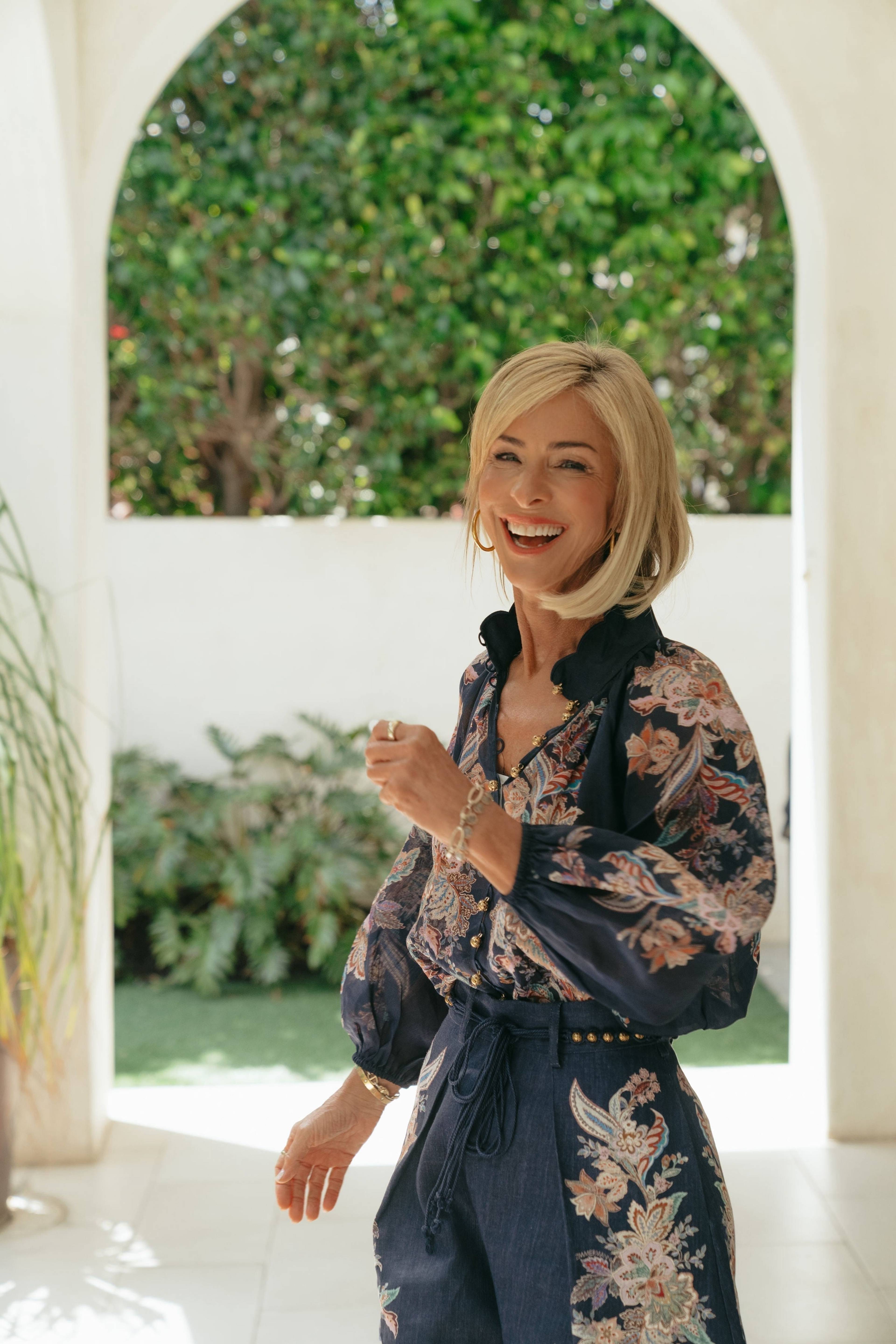 A smiling blonde woman in a dark floral-patterned outfit stands beside an arched opening with greenery behind her.