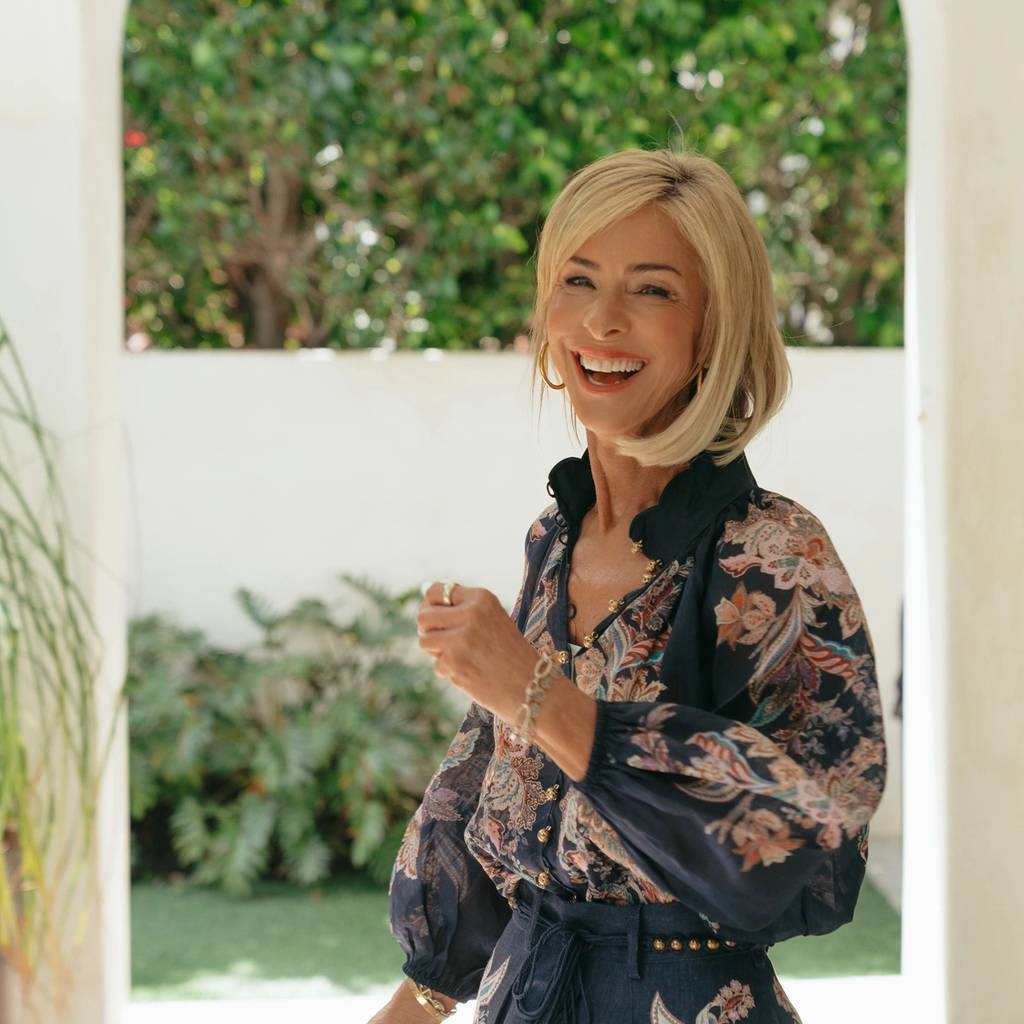 A smiling blonde woman in a dark floral-patterned outfit stands beside an arched opening with greenery behind her.