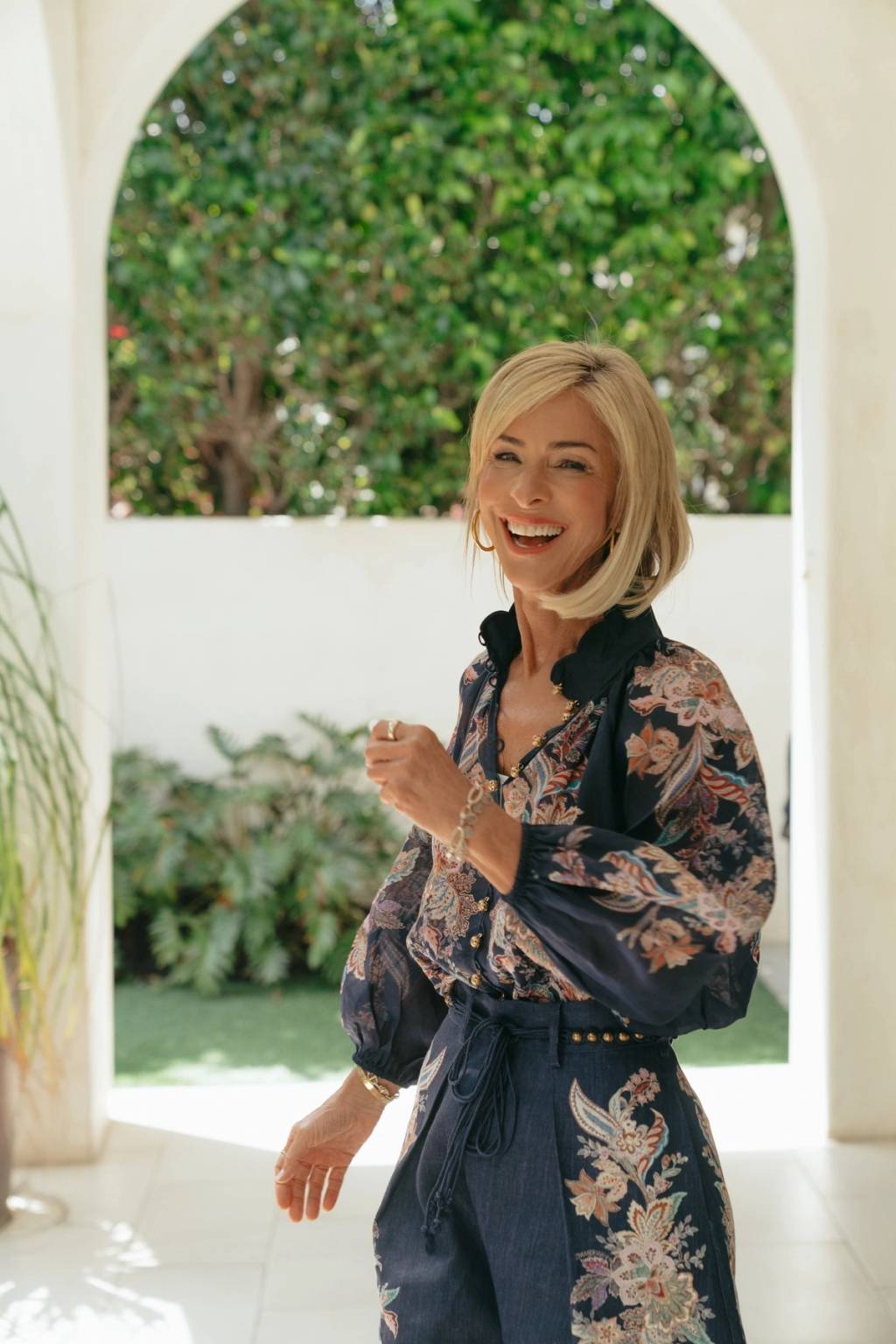 A smiling blonde woman in a dark floral-patterned outfit stands beside an arched opening with greenery behind her.