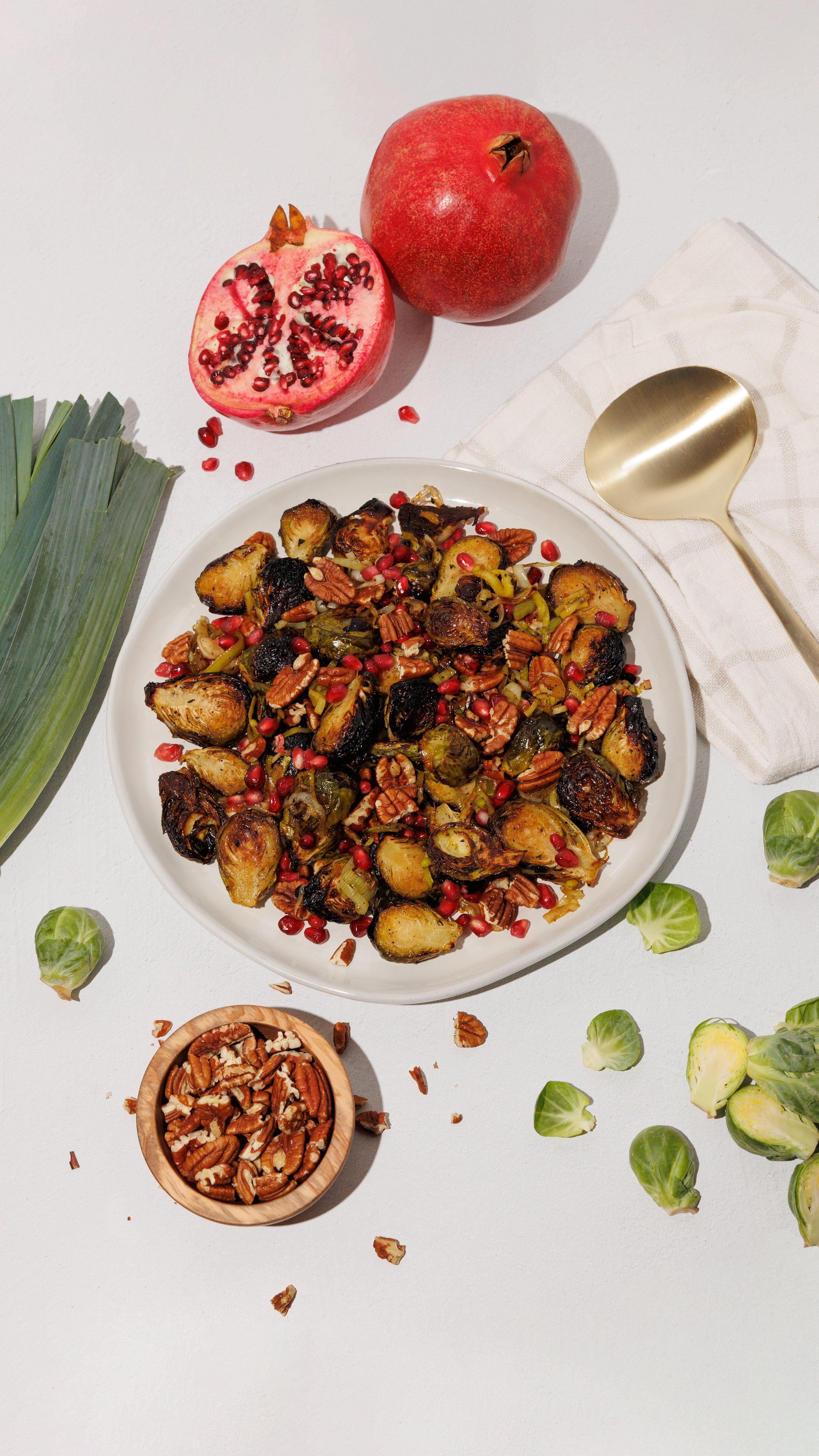 Plate of roasted Brussels sprouts with pomegranate seeds and pecans, surrounded by fresh ingredients.
