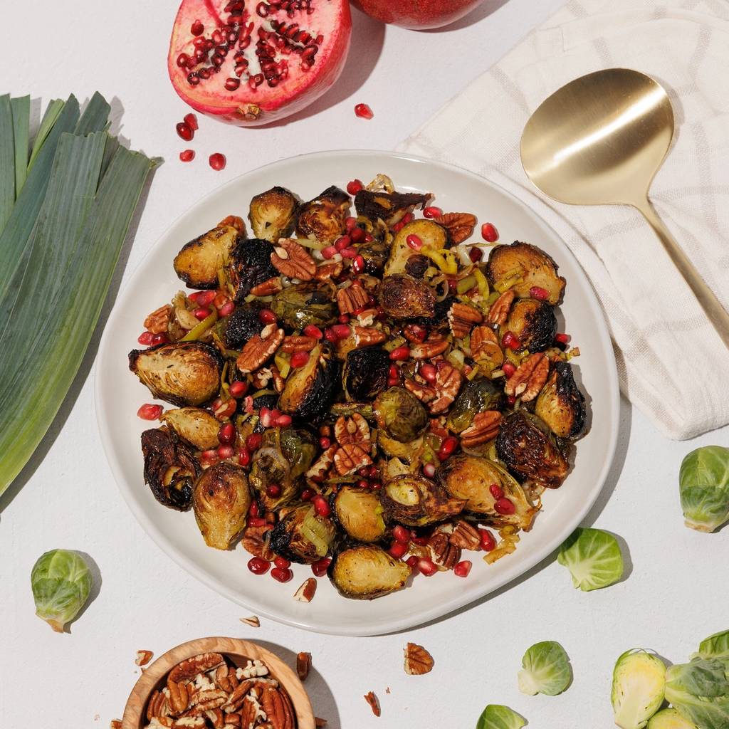 Plate of roasted Brussels sprouts with pomegranate seeds and pecans, surrounded by fresh ingredients.