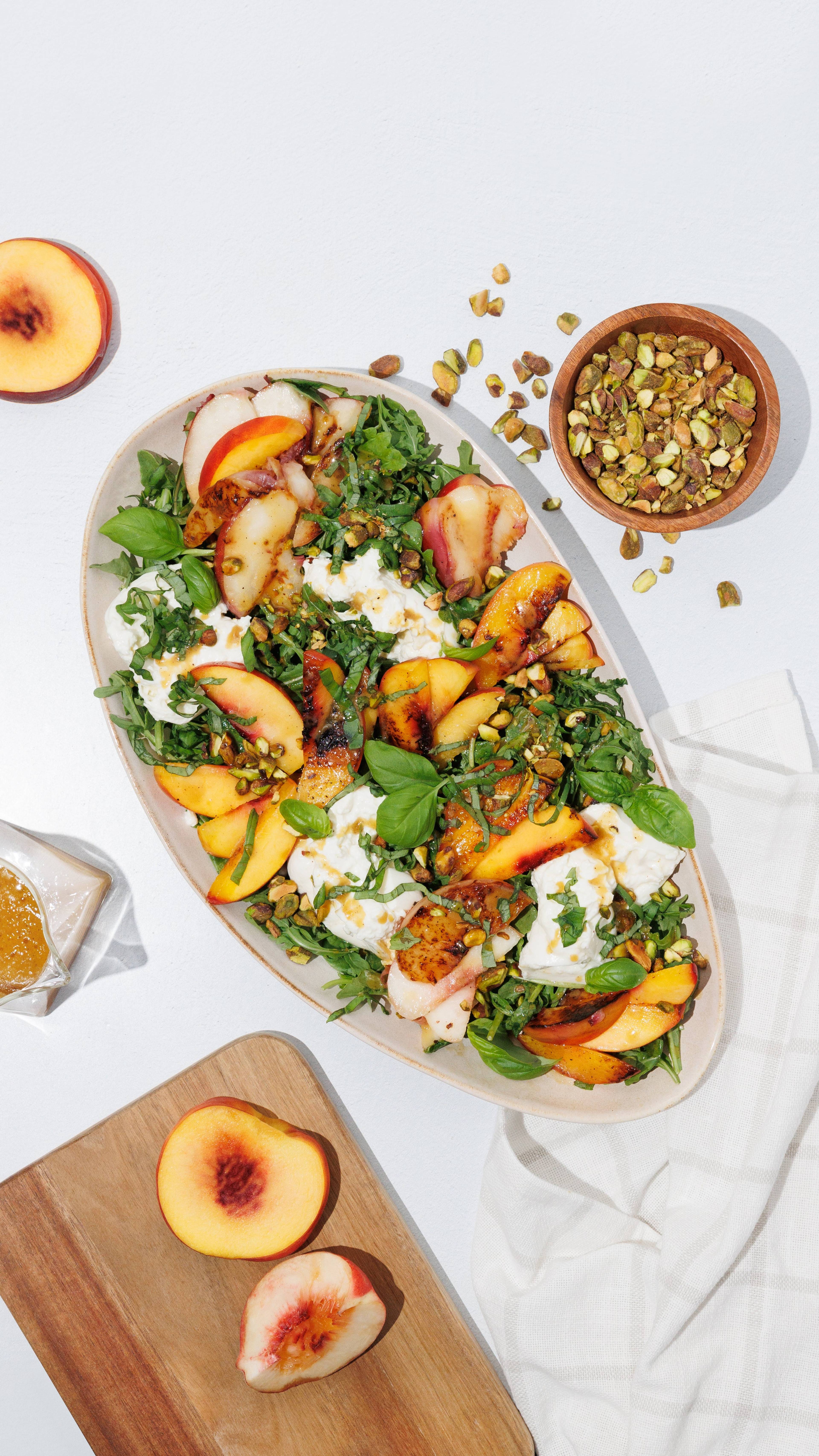Platter of peach, burrata, arugula, basil, and pistachio salad with halved peaches and honey nearby.