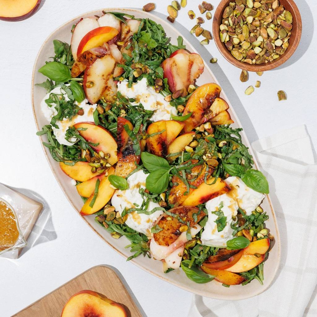 Platter of peach, burrata, arugula, basil, and pistachio salad with halved peaches and honey nearby.