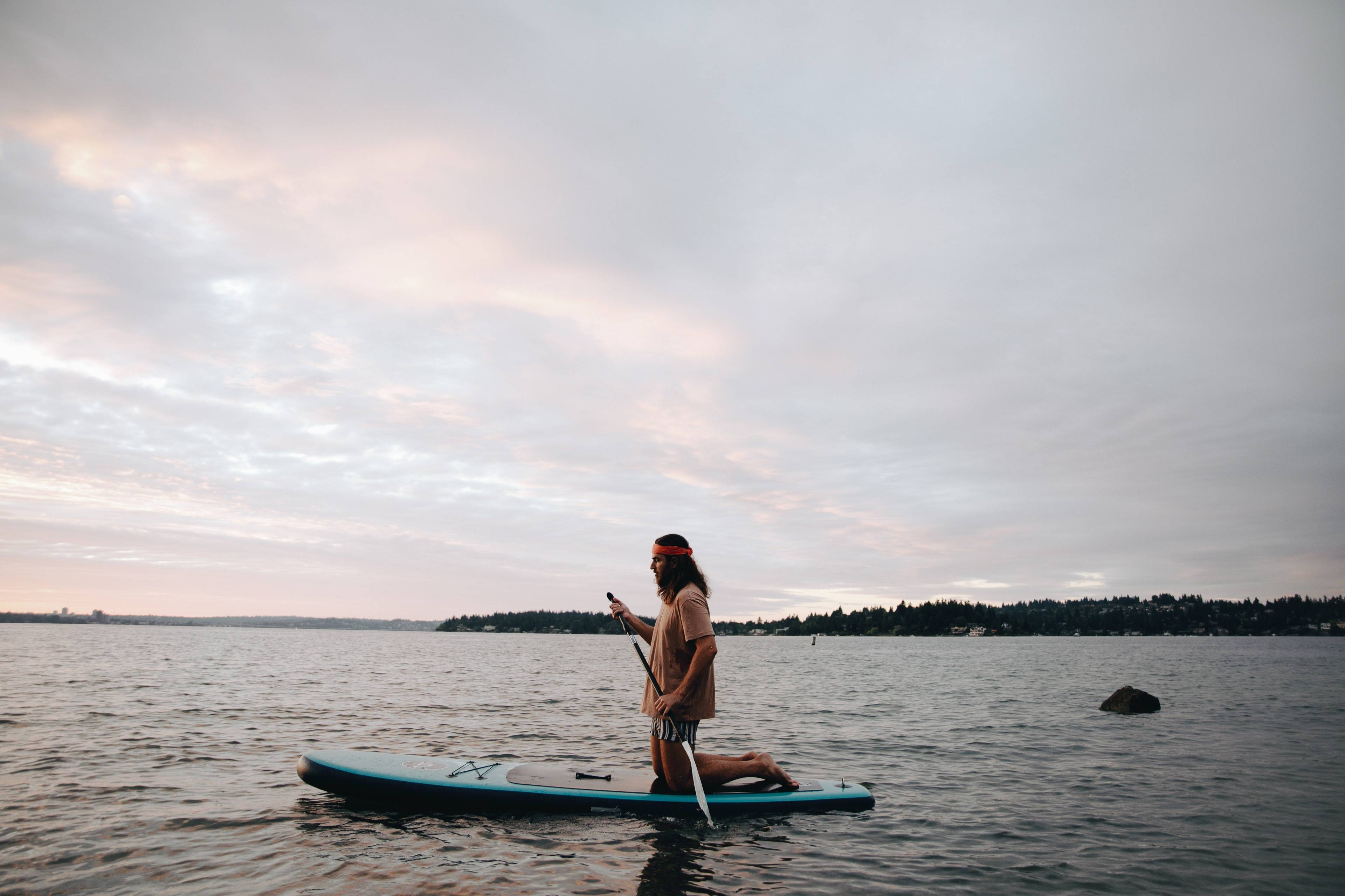 Person kneeling on a paddleboard and holding a paddle on calm water under a cloudy sky.