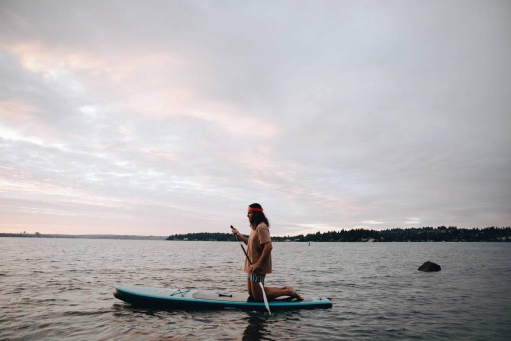 Person kneeling on a paddleboard and holding a paddle on calm water under a cloudy sky.