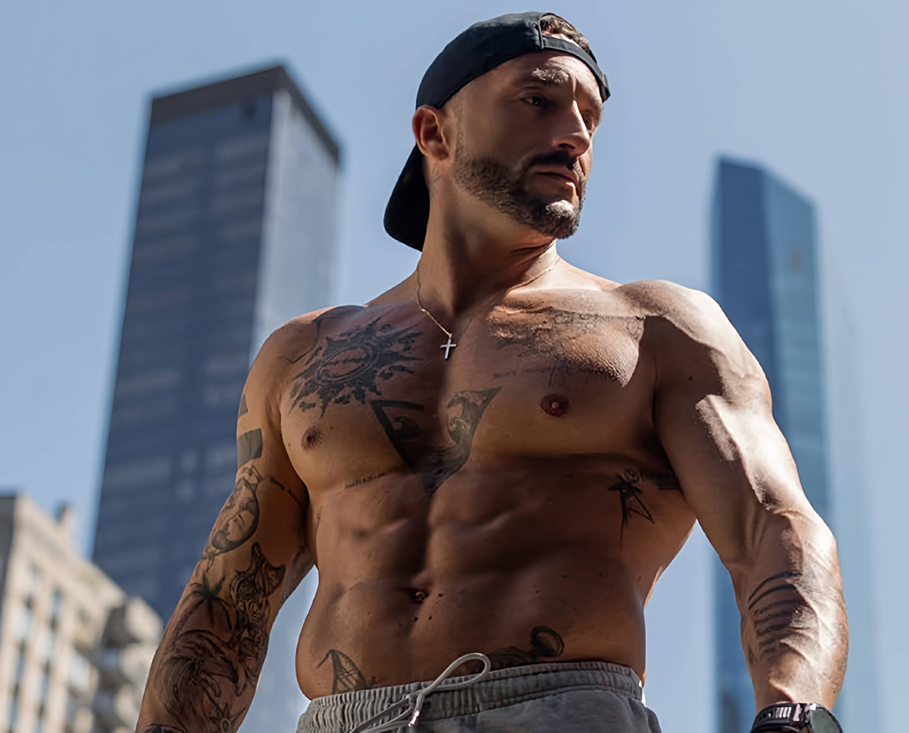 Shirtless tattooed man in a backward cap standing outdoors between tall city buildings.