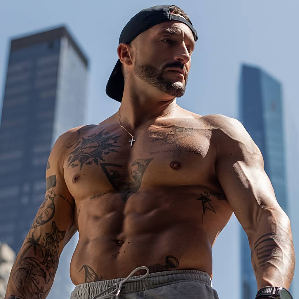 Shirtless tattooed man in a backward cap standing outdoors between tall city buildings.