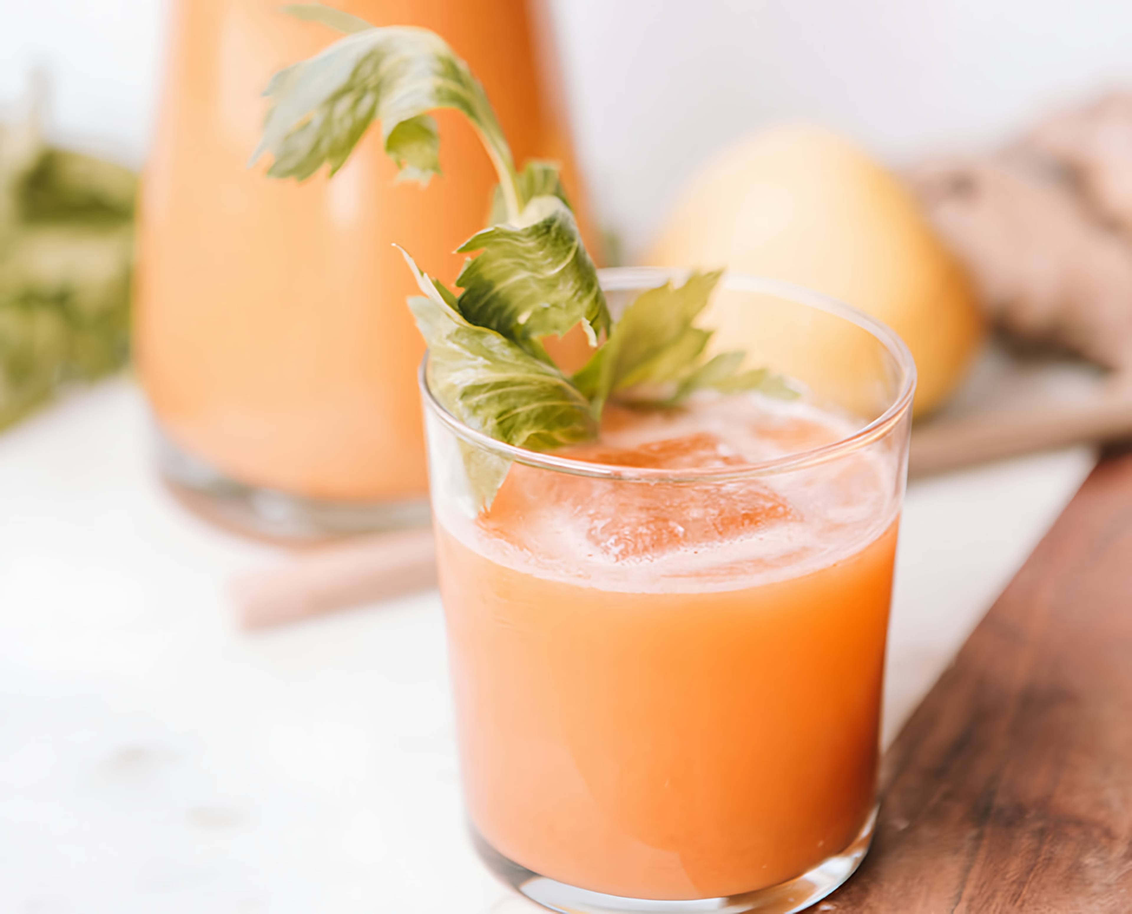 Glass of orange juice with ice and leafy garnish, with a pitcher of juice and a lemon in the background.