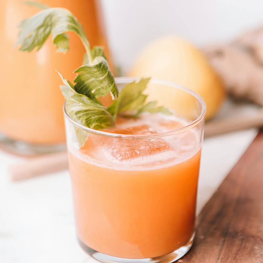 Glass of orange juice with ice and leafy garnish, with a pitcher of juice and a lemon in the background.