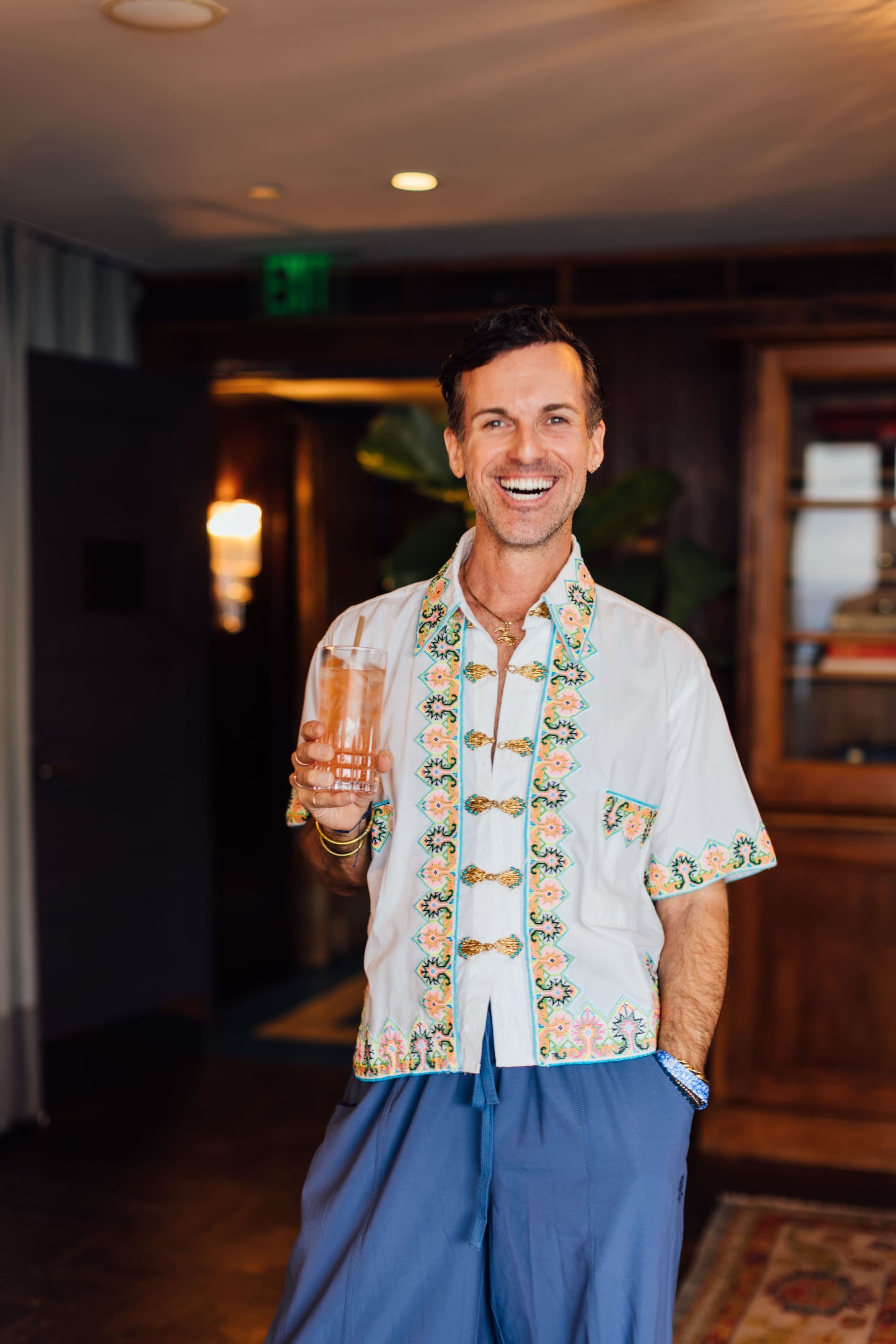 Smiling man in an embroidered white shirt holding a glass indoors.