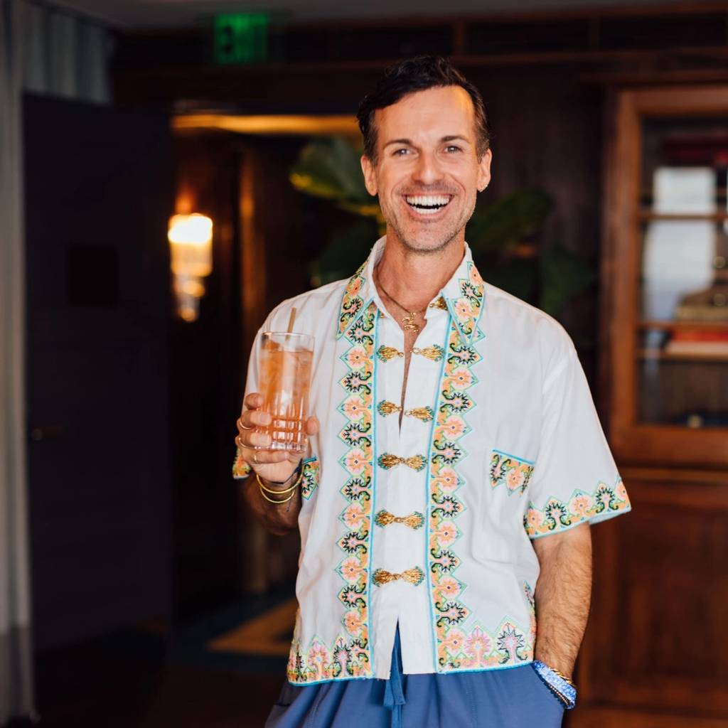 Smiling man in an embroidered white shirt holding a glass indoors.